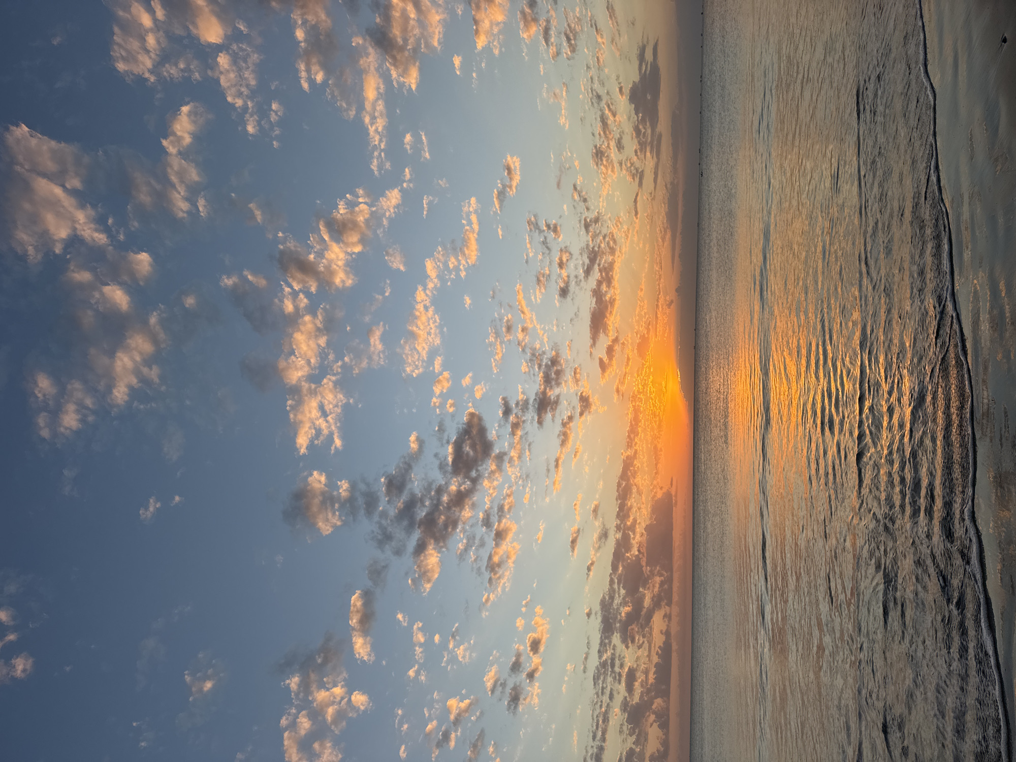 The image captures a tranquil seascape at sunset, viewed from a sandy beach. The sun, a vibrant orange disk, is partially obscured by the horizon, casting a warm, golden glow across the sky and the calm ocean surface. Scattered clouds, tinged with pink and orange hues, dot the vast expanse of the blue sky. The water, reflecting the sunset colors, exhibits gentle ripples. In the distance, several small boats are visible on the water, suggesting a coastal area. The foreground shows the wet sand of the beach with a thin line of foam from receding waves. The overall impression is one of peace and natural beauty, characteristic of a serene evening in Kuta Selatan, Indonesia, a well-known tourist destination for its beaches and sunsets.