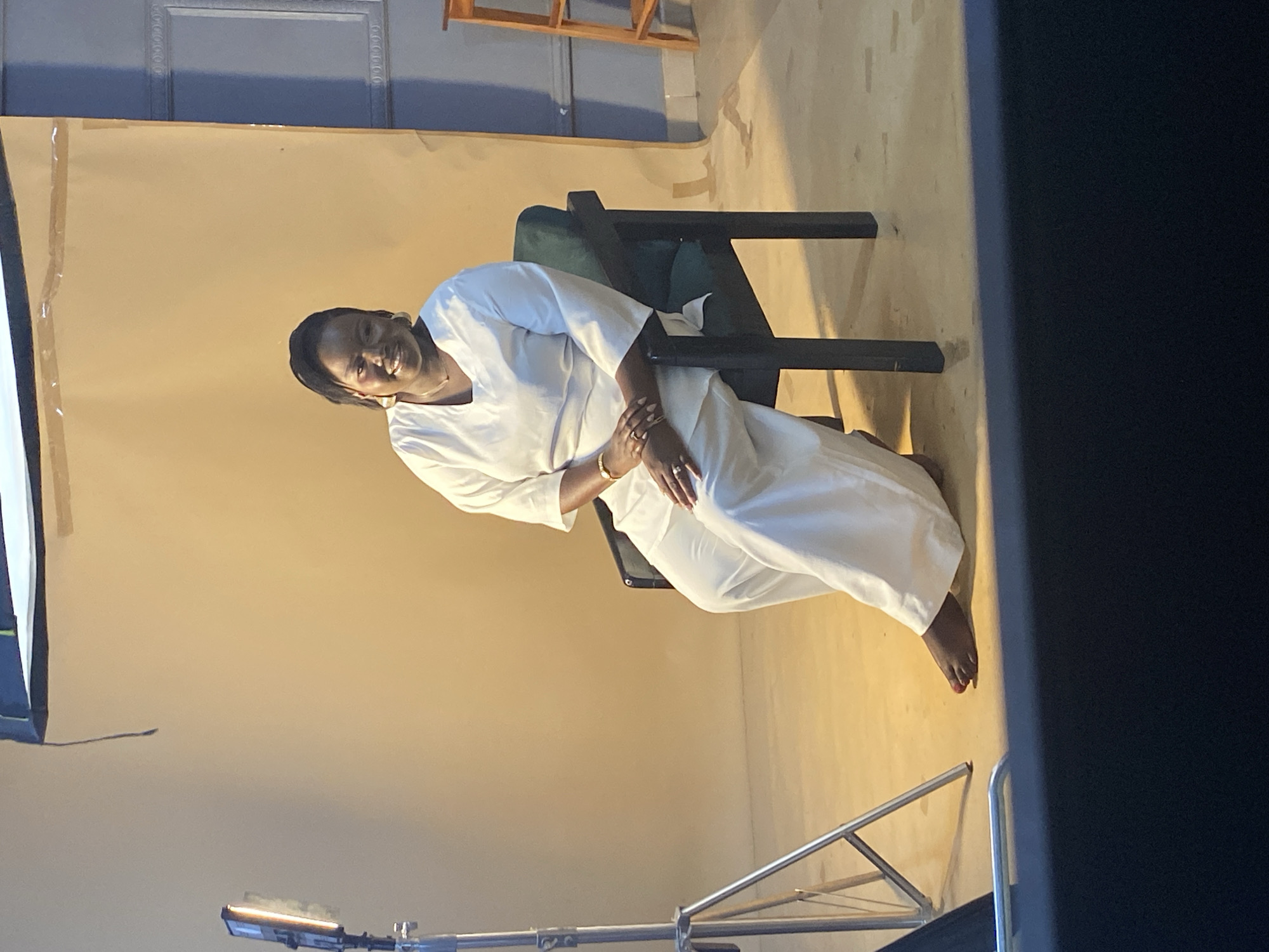 A woman is seated in a chair, wearing a white outfit. She is looking towards the camera and smiling. The background appears to be a studio setting, with a plain, light-colored backdrop and some lighting equipment visible in the foreground. The scene is lit with artificial light, suggesting it is indoors or at night. The woman has dark skin and dark hair, and is wearing earrings and rings on her fingers. The overall mood is relaxed and friendly. There is no visible text in the image.