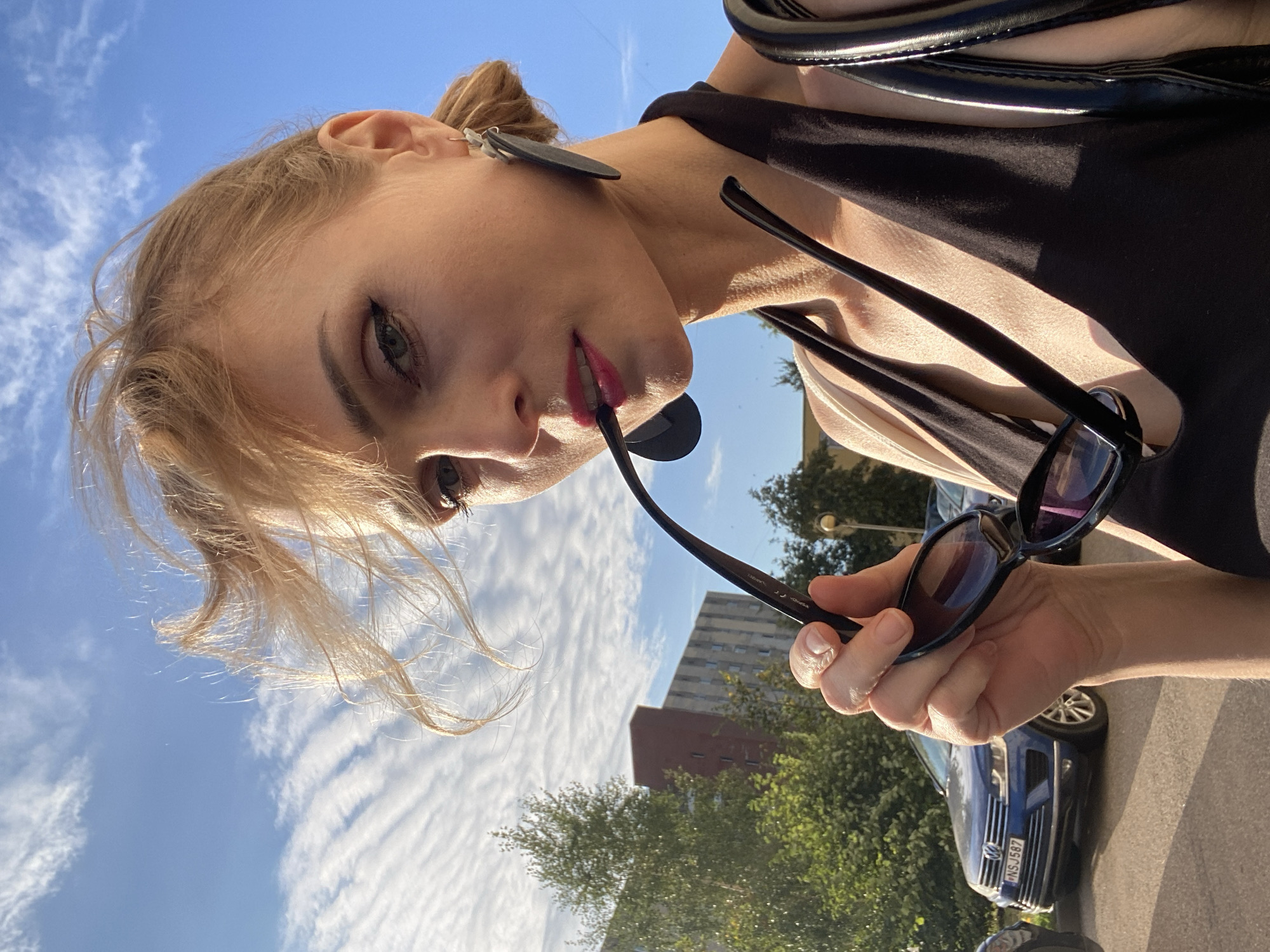 A woman with blonde hair styled loosely and windblown, wearing dark sunglasses, and dark lipstick, holds the sunglasses up to her mouth as she looks at the camera. She is wearing a black halter top and large dark earrings. In the background, a blue Volkswagen SUV is parked in a lot. A building with a red top is visible behind the SUV, as well as trees and a blue sky with wispy white clouds. The image was taken in Lida, Belarus.