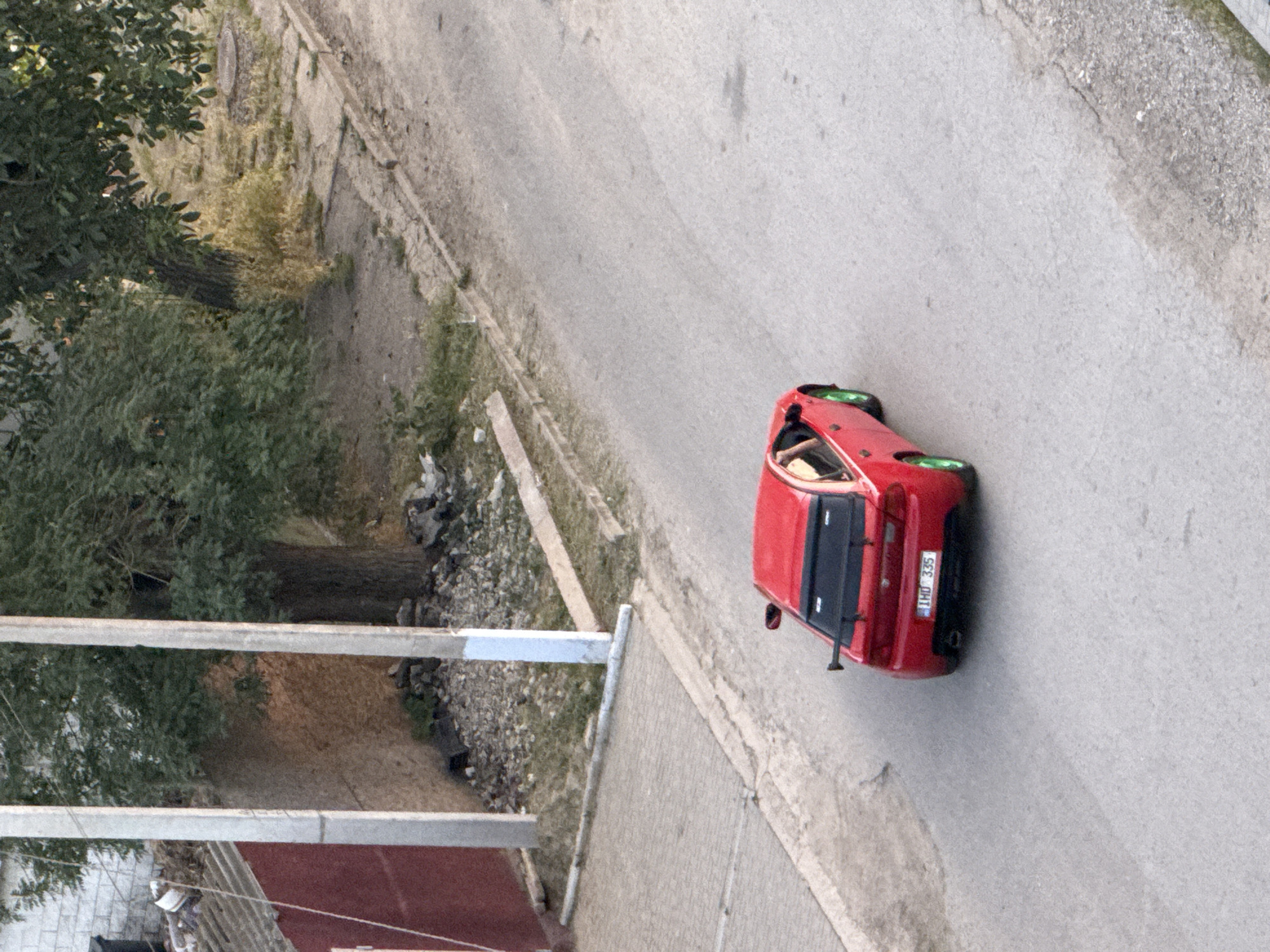 A red sedan with green rims is driving on a paved road. The car has a spoiler on the back and the license plate reads "IHD 335". A person's arm is visible from the driver's side window. The road is bordered by a sidewalk and a grassy area with trees. To the left, there is a red building and some debris. The image appears to be taken from an elevated position, looking down on the street. The scene is consistent with a residential area in the city of Cahul, Moldova, likely during daylight hours.