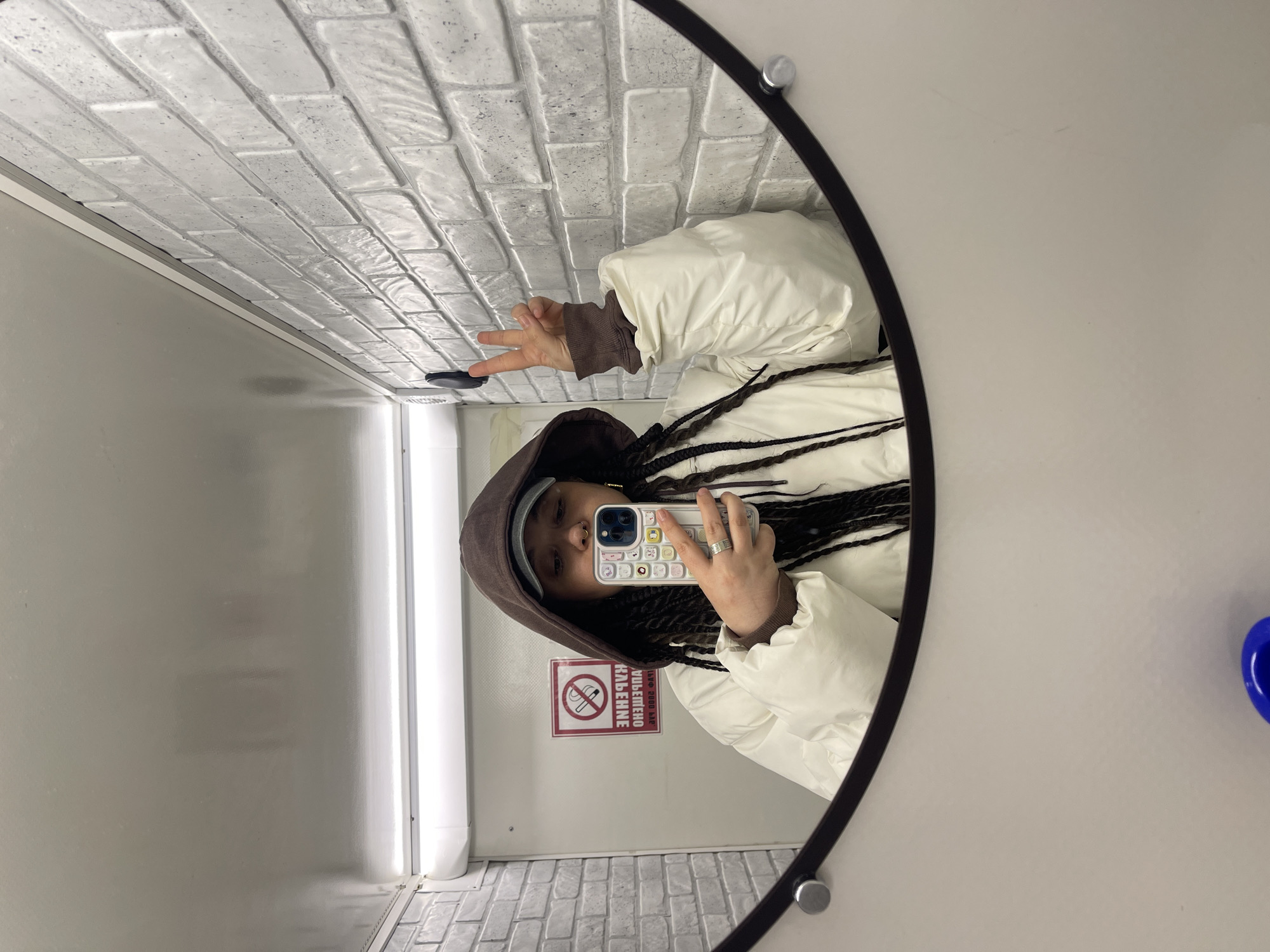 A person in a brown hoodie, white jacket, and braids is taking a selfie in a mirror. The mirror reflects a bathroom stall with white brick-patterned walls, a fluorescent light, and a "no smoking" sign in Russian, suggesting the location is likely in Russia, possibly Moscow. The person is holding up two fingers in a "peace sign."