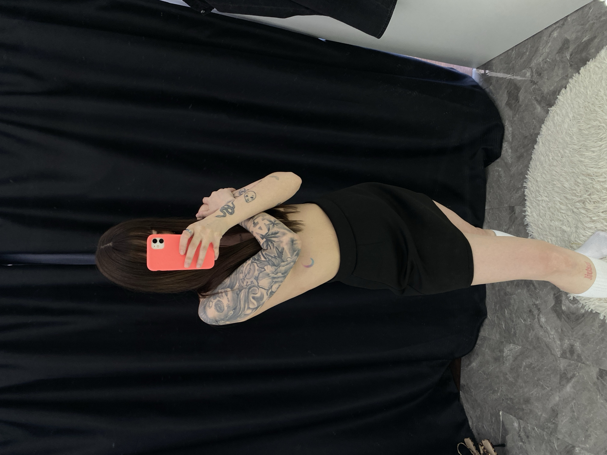 A top-down, eye-level photograph depicts a person with brown hair, lying on a black fabric surface with their back exposed. They are holding an orange mobile phone in their hands, covering part of their face. Visible tattoos are present on the person's arms. The subject is wearing a black garment that covers part of their lower torso and legs. The person is positioned near a white fluffy rug, which sits on a gray-toned, marbled surface. The location in Izhevsk, Russia cannot be determined from this image.