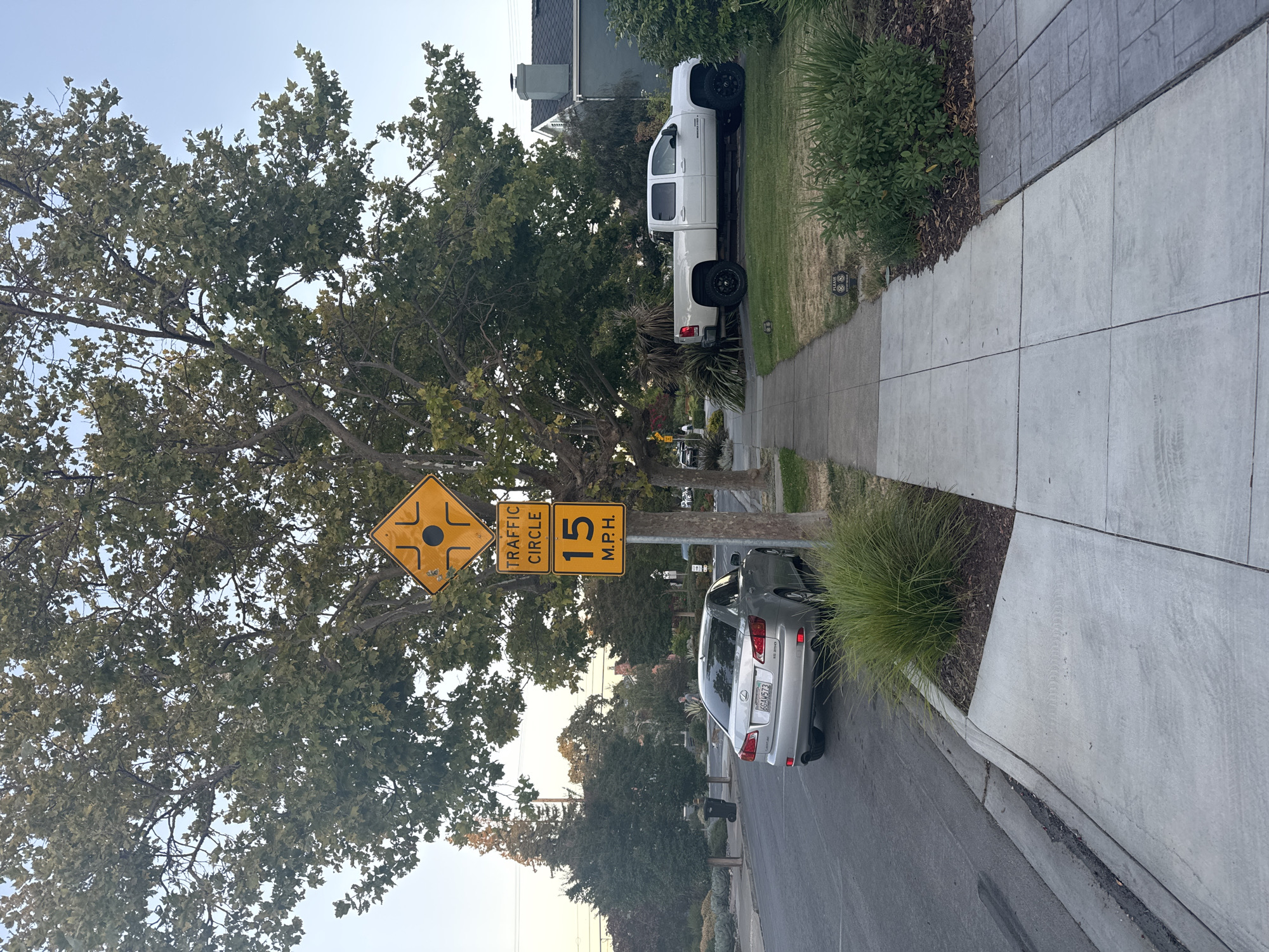The image depicts a residential street scene in San Carlos, United States of America. A silver Lexus sedan is parked on the left side of the street, facing away from the camera. A white pickup truck is parked on the right side of the street, also facing away from the camera. Two traffic signs are visible on a pole on the right side of the image. The top sign is a yellow diamond with black symbols indicating a traffic circle. The bottom sign is also yellow and states "TRAFFIC CIRCLE 15 M.P.H.". Trees with green leaves are present on both sides of the street, and a sidewalk runs along the right side. Houses and other residential structures are visible in the background. The sky is clear and blue. The lighting suggests it is either early morning or late afternoon.