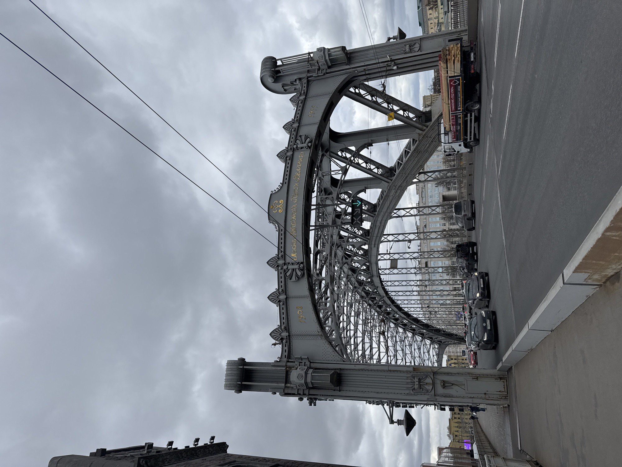 The image displays a large, ornate metal truss bridge under a heavily overcast, grey sky. The perspective is from below and to the side, highlighting the intricate grey metallic structure composed of riveted arches, lattice work, and decorative elements. Golden Cyrillic script and the year "1908" are prominently inscribed on the main arch of the bridge, identifying it as the "Bridge of Emperor Alexander III."
The top of the bridge features a paved roadway with multiple lanes of traffic. Several dark-colored cars are visible, alongside a white truck carrying a load of wooden planks or lumber. The truck also bears Cyrillic text on its side, indicating "House Renovation - Cut Materials." Overhead power lines stretch across the sky in the upper left portion of the image.
Multi-story buildings with classic European architecture can be seen in the background, lining the banks below and beyond the bridge. The lighting is flat due to the cloudy conditions, suggesting daytime without direct sunlight. The scene depicts an active urban environment, characteristic of Saint Petersburg, Russia, with the historic bridge serving as a central element of daily life and transportation.