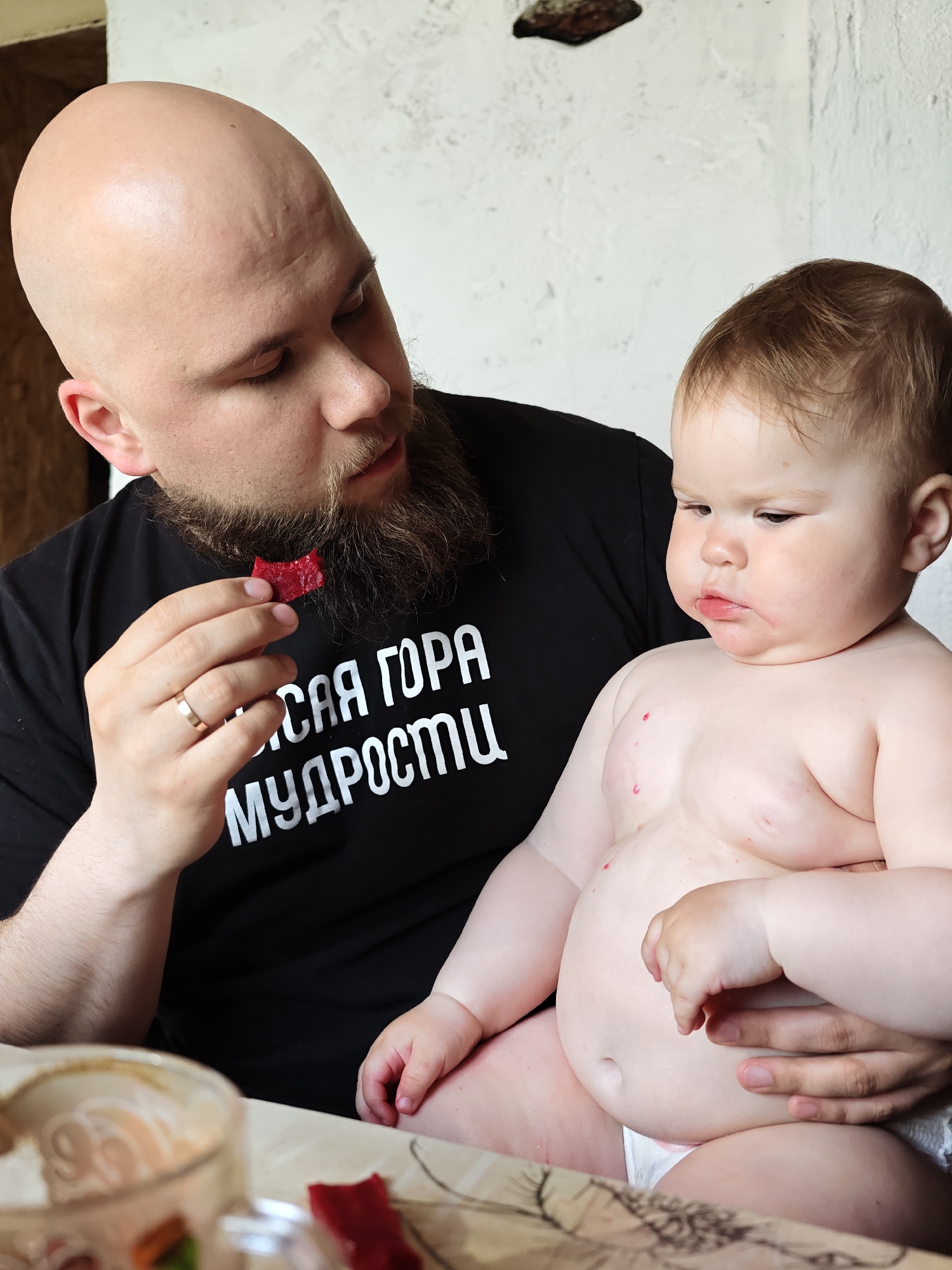 The image shows a bald, bearded man wearing a black t-shirt with white Cyrillic text holding a young, shirtless child. The man is holding a small, red piece of food in his fingers and appears to be offering it to the child, who looks away with a sullen expression. The child has several red spots on their chest and arms. A table with a partly-eaten dish sits in the foreground. The location, Malyi Bukor, Russia, is not identifiable from the image. No further details about the individuals, actions, or environment can be determined from the image.
