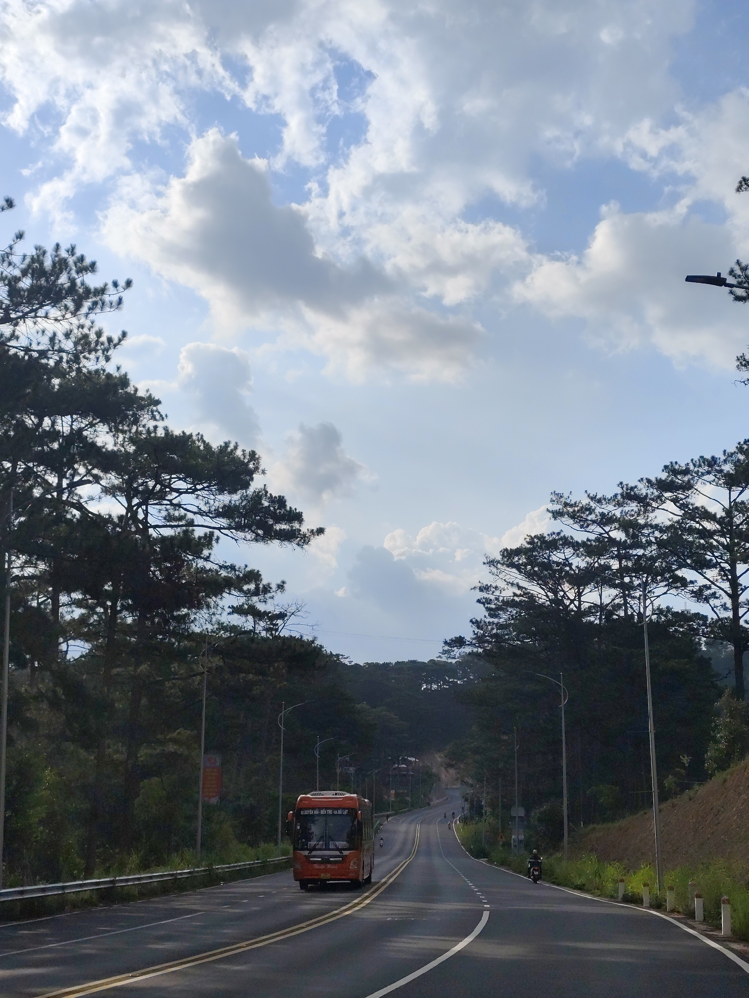 A low-angle outdoor shot depicts a roadway running through a forested area under a partly cloudy sky. The road, marked with double yellow lines, curves gently into the distance, leading toward a low mountain ridge. An orange bus is traveling on the road towards the camera. In the distance, a motorcycle is traveling on the roadway. Tall pine trees line both sides of the road. Streetlights are positioned along the roadway. The location is Da Lat, Vietnam.
