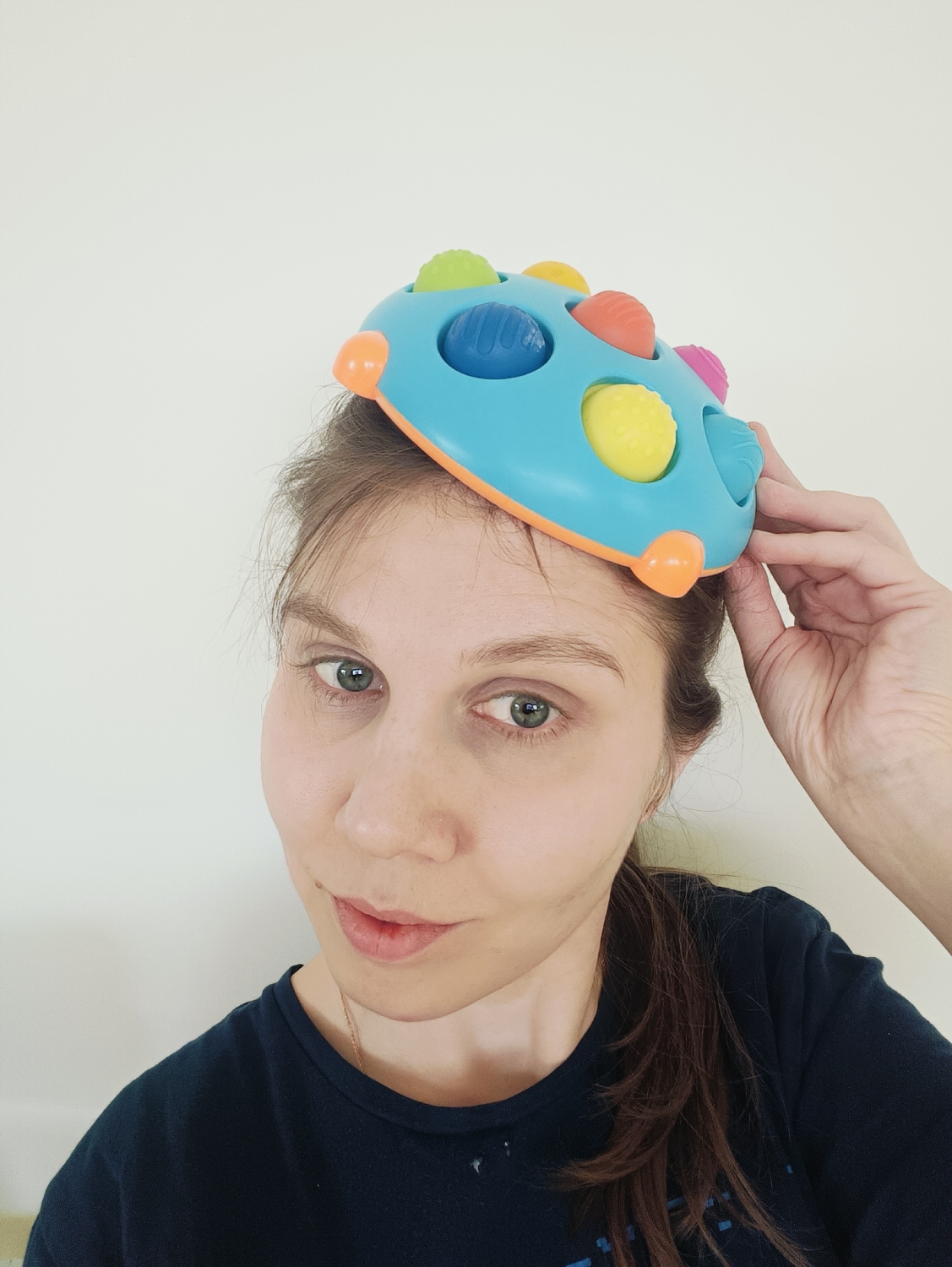 A young to middle-aged woman is depicted in a close-up, head-and-shoulders portrait, likely a selfie, against a plain, light-colored wall, indicating an indoor setting. She has light skin, green eyes, and brown hair pulled back into a ponytail. She wears a dark blue t-shirt. A colorful fidget toy rests on top of her head, with her left hand lightly holding it in place on the right side of the frame. The toy is light blue and oval-shaped with an orange border, and features multiple textured pop-it bubbles in yellow, green, blue, orange, and pink. Some of these bubbles are depressed. The woman looks directly at the camera with a neutral to slightly playful expression, characterized by a subtle smirk on her lips. The lighting is bright and even, suggesting daytime. There is no visible text in the image.