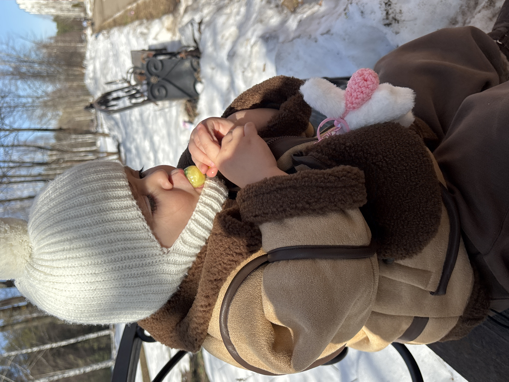 A young child is seated on a black metal bench in Chaikovsky, Russia. The child is wearing a white knitted hat with a pom-pom, a tan and dark brown shearling-style coat, and dark brown trousers. They are holding a yellow-green lollipop in both hands, bringing it to their mouth. A white and pink stuffed animal keychain is clipped to the child's coat. The surrounding environment consists of a paved pathway running alongside the bench, with melting snow and patches of brown earth. Bare deciduous trees and some coniferous trees are visible in the background, with a multi-story building in the far distance under a clear blue sky.
