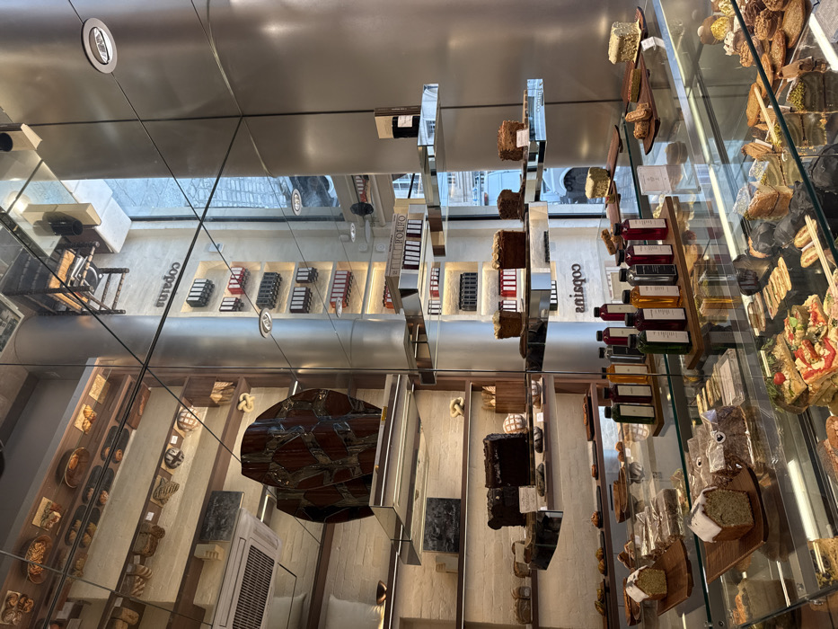 The image captures the interior of Copains, a modern bakery located at 18 Rue Bachaumont, 75002 Paris, France. The scene emphasizes a sleek, contemporary design with extensive use of reflective surfaces, including mirrored walls, glass display cases, and polished metal accents.
In the foreground, a multi-tiered glass display case showcases a diverse array of pastries, cakes, and savory items. Visible offerings include individual cake slices, cookies, tarts, and small rectangular pizzas topped with vegetables. Above this display, on wooden boards, several loaf cakes are presented, some adorned with white icing. Adjacent to these are rows of bottled beverages in various colors (e.g., dark green, red, orange, clear), arranged on wooden trays.
Mid-ground elements include larger rectangular cakes, such as a dark chocolate cake, positioned on mirrored shelves. A prominent, dark brown sculptural object with a fragmented, geometric pattern rests on a mirrored pedestal on the left. Reflections in the mirrored surfaces prominently display the inverted name "copains."
The background features shelves against a light-colored wall, displaying various baked goods like bread and pretzels. Another section of the wall holds multiple rows of dark, rectangular packaged items. Large windows reflect portions of the exterior, showing a paved street or cobblestone area and architectural details of an opposing building. Recessed lighting fixtures illuminate the space, and an air conditioning vent is visible on the upper left wall. No individuals are present within the scene.