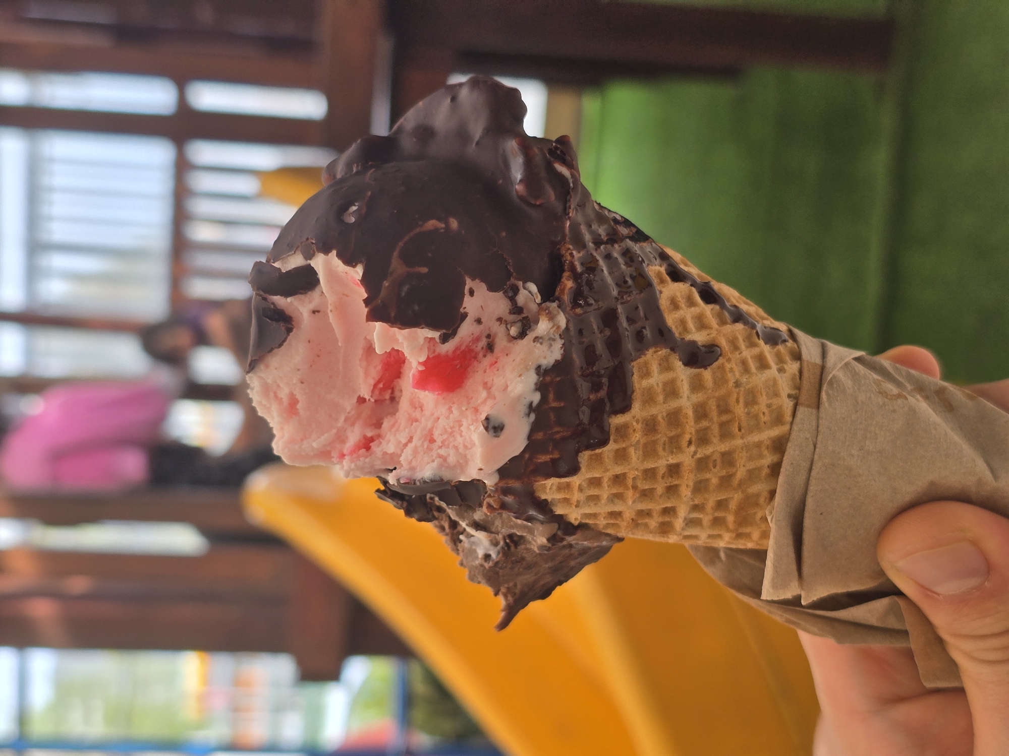 A close-up shot shows a hand holding a partially eaten ice cream cone. The ice cream is pink with red flecks, covered in a layer of dark chocolate. The waffle cone is wrapped in a brown paper wrapper. The background is blurred, but it appears to be a playground with a yellow slide and various wooden structures. There are indistinct figures in the background. The location is possibly a playground in Puente Alto, Chile.