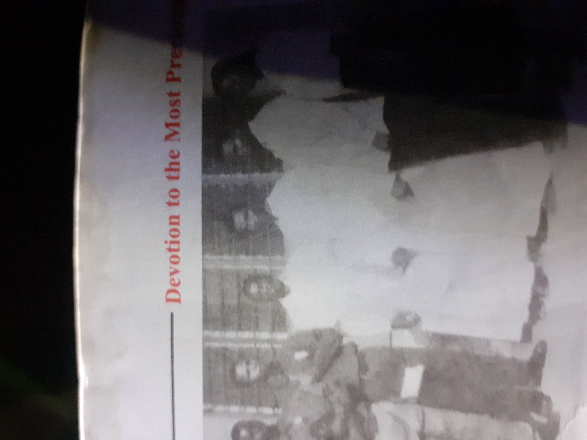 The image displays a photograph featuring a group of people in front of a building. Text above the individuals reads "Devotion to the Most Precious". The individuals appear to be men, some are wearing uniforms, and others are in what looks like religious or ceremonial attire. The building behind them has visible windows. The lighting is uneven, with some areas of the image being significantly darker than others, suggesting it might be an indoor photograph taken with flash or an outdoor photo with challenging lighting conditions. The overall image quality is poor due to blur and low resolution, making it difficult to discern fine details about the individuals' facial features or the specifics of their clothing and the environment. No specific location in the city of Jalingo, Nigeria can be determined from the visual information present.