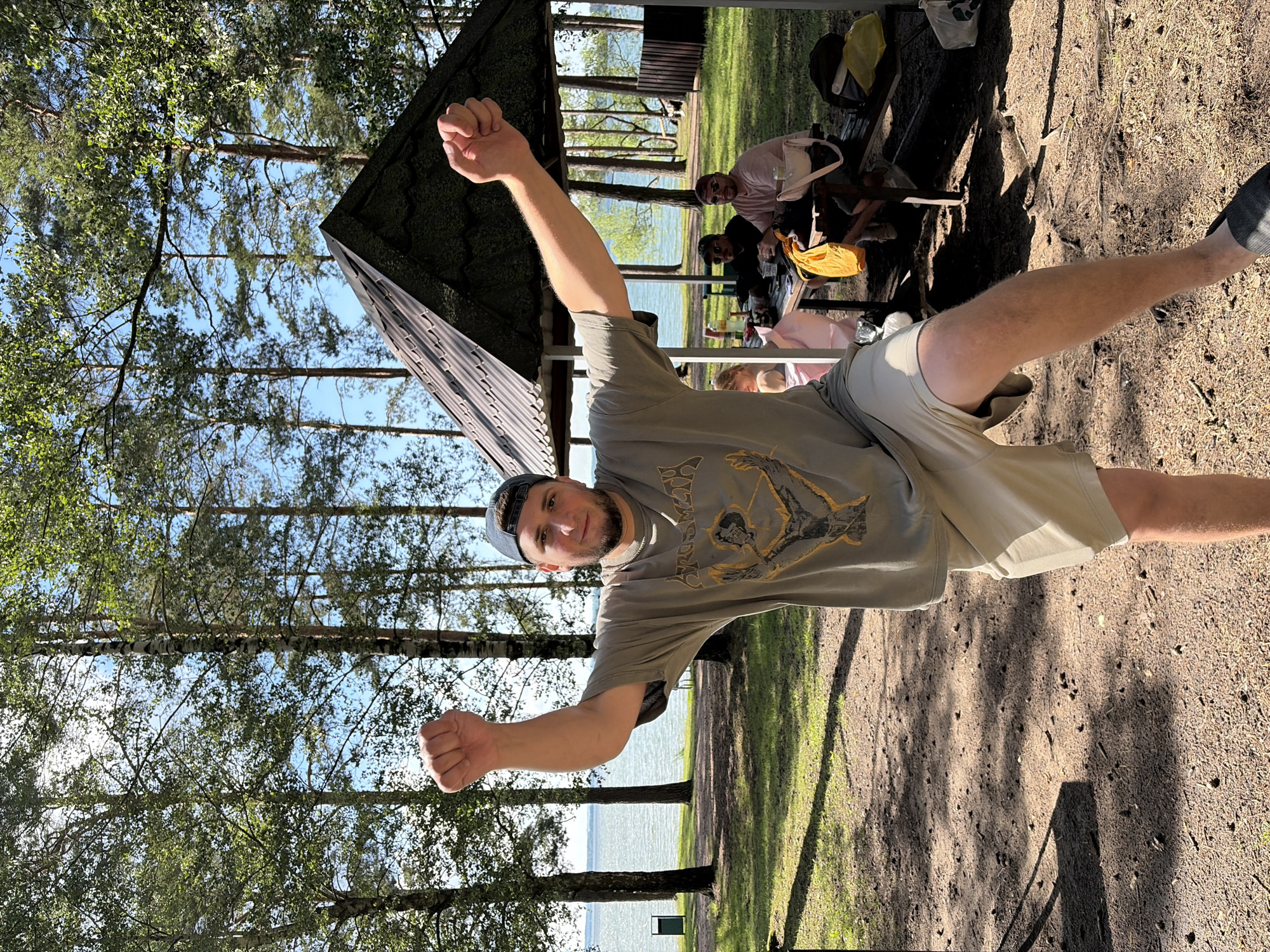A man, wearing a grey t-shirt with a graphic design, tan shorts, a dark baseball cap, and grey socks with black shoes, poses with his fists clenched and arms extended in a park-like setting. He is facing the camera and appears to be smiling. In the background, other people are visible sitting at a picnic table under a covered structure. There is a body of water, likely a lake, and trees surrounding the area. The sun appears to be shining, casting shadows on the ground. The overall scene suggests a relaxed outdoor gathering. No visible text is present in the image.