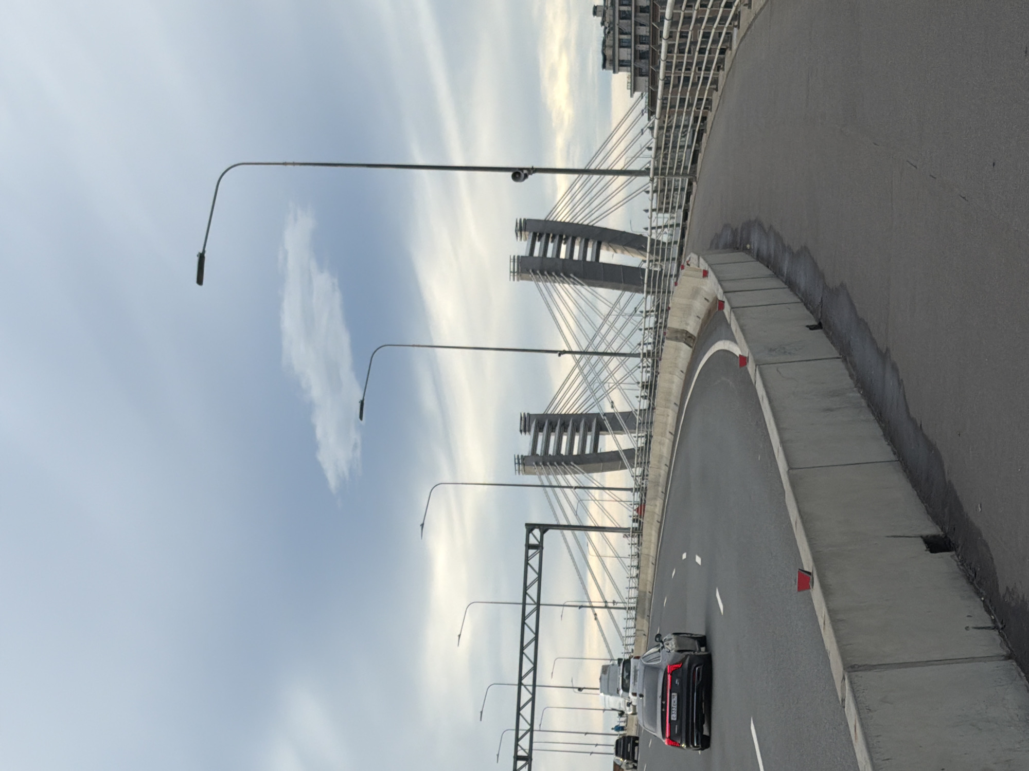 The image shows a view from a car driving on a bridge in Saint Petersburg, Russia. The bridge has a modern design with suspension cables and tall concrete pillars. The road is asphalt and has lane markings. Streetlights line the side of the road.
In the foreground, a black Lexus is driving on the bridge. The car has its brake lights on, indicating it is either slowing down or stopped. Other vehicles are visible in the distance. The sky is overcast with streaks of white clouds. The time of day appears to be late afternoon or early evening, given the soft light.
The background shows a cityscape with buildings, but they are out of focus. The overall scene is urban and functional, typical of a bridge on a busy road. There is no visible text in the image.