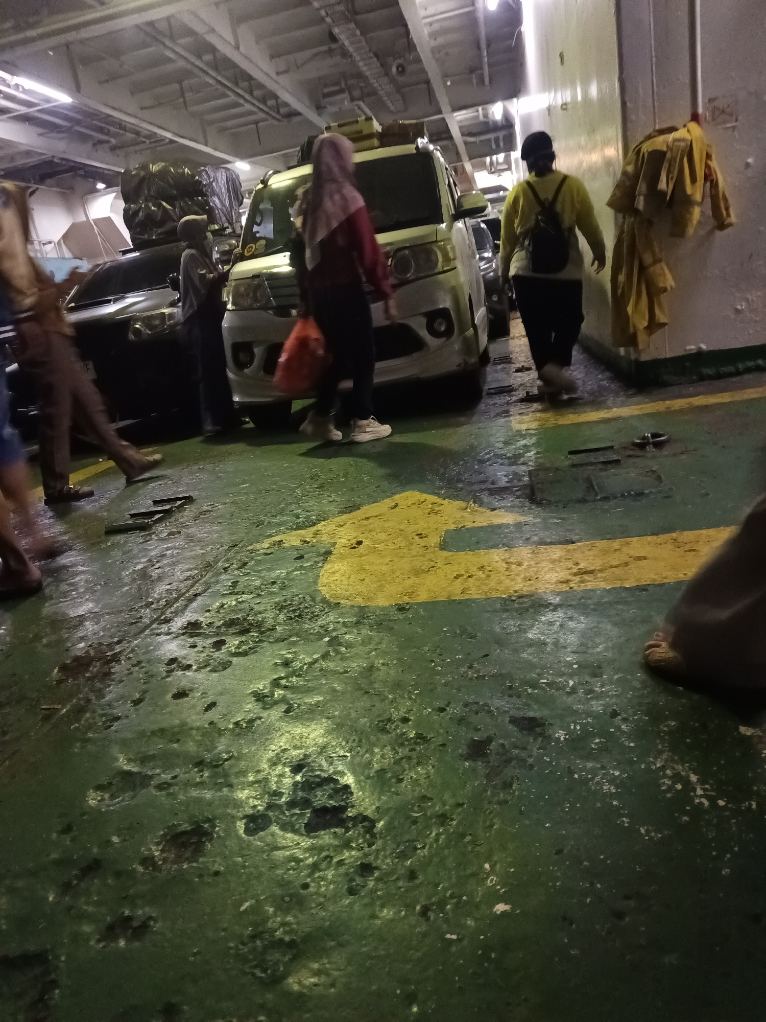 The image depicts the interior of a ferry in Cilegon, Indonesia, likely transporting vehicles and passengers. In the foreground, the wet, green, and yellow painted deck shows signs of wear and tear. Several people are visible, some walking and others standing near vehicles. A light-colored van is parked in the middle ground, with a person in a maroon headscarf and jacket carrying a red bag walking past it. To the left, part of a black car is visible. Further to the right, a person wearing a yellow jacket, black pants, and a backpack is walking away from the camera. Yellow jackets are hanging on a wall to the right. The scene is dimly lit, suggesting it's either inside the ferry's vehicle deck or during low light conditions.