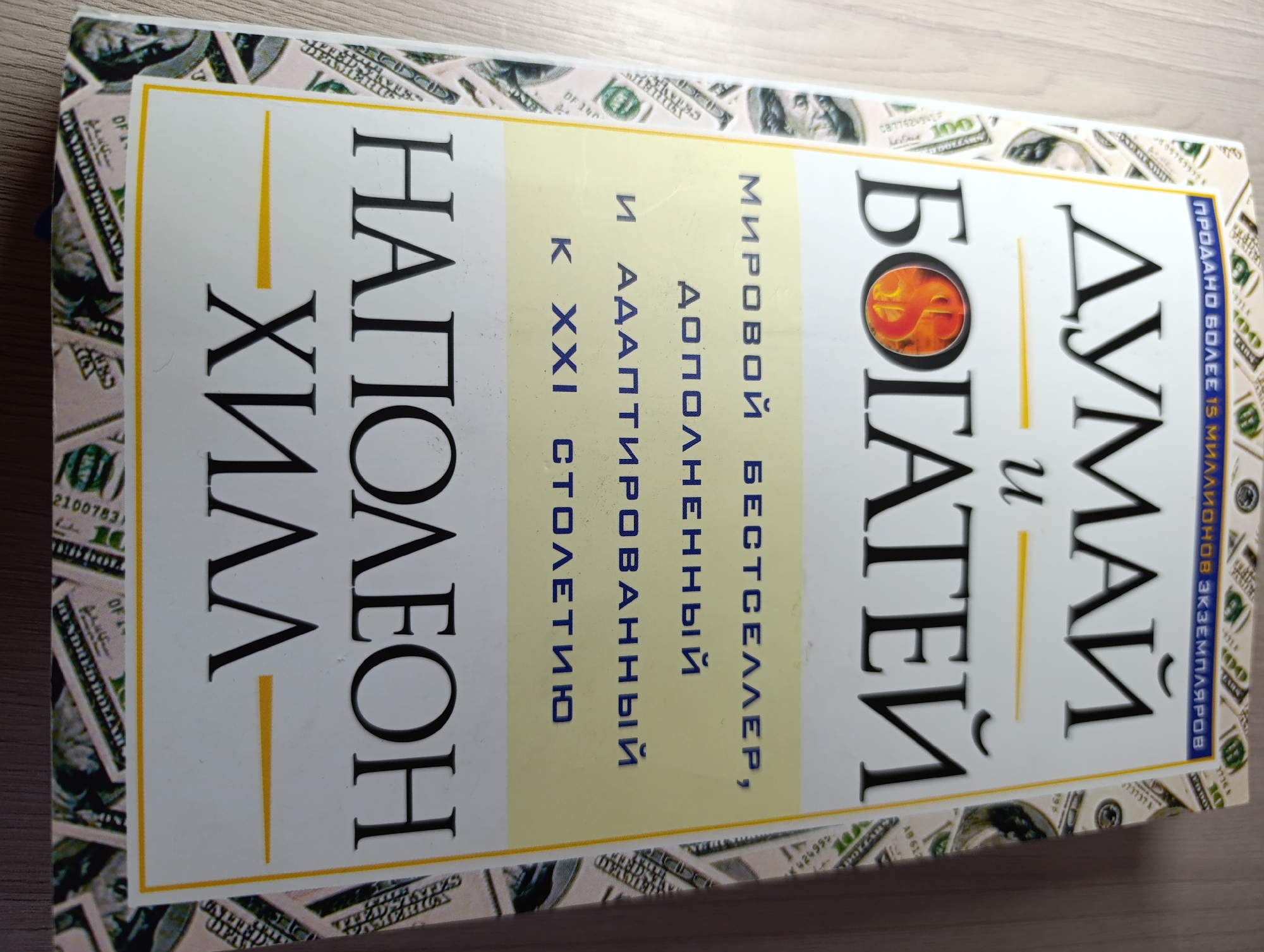 The image shows the cover of a book titled "Думай и Богатей" (Think and Grow Rich) by Napoleon Hill. The book cover is white with a yellow border and features large black lettering. The title is prominent, with "ДУМАЙ" on the right and "и БОГАТЕЙ" on the left, separated by a vertical yellow line. Below "БОГАТЕЙ" is a graphic of a dollar sign within a flame. To the left of this, in a vertical orientation, is "НАПОЛЕОН ХИЛЛ". Smaller text is also visible, including "МИРОВОЙ БЕСТСЕЛЛЕР" (World Bestseller) and "АДАПТИРОВАННЫЙ К XXI СТОЛЕТИЮ" (Adapted for the XXI Century). The book cover is placed on a wooden surface and is surrounded by imagery of US dollar bills. The scene is an indoor, studio-like setting with no visible city or environmental context.
