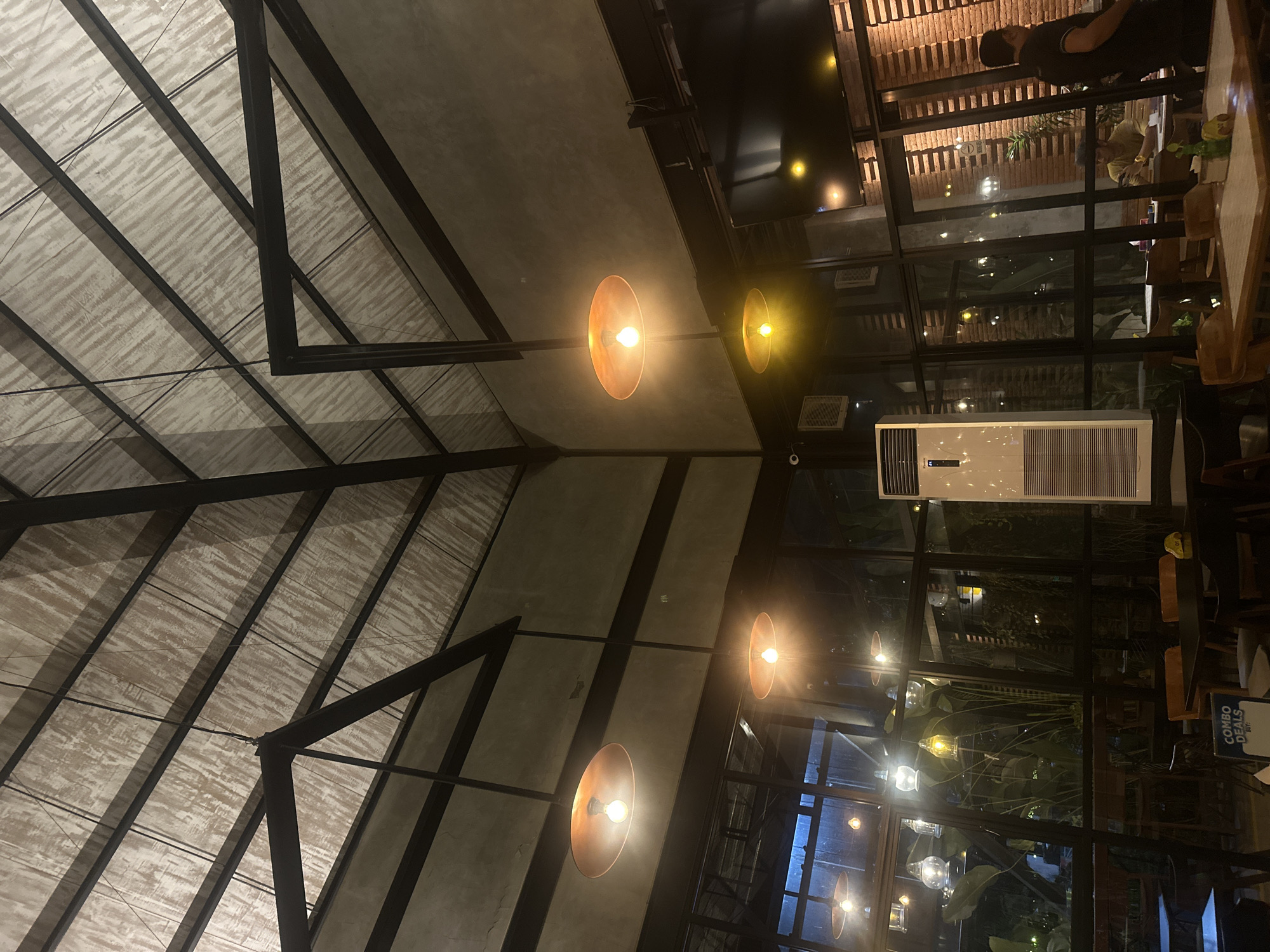 The image depicts the interior of a restaurant with a high, vaulted ceiling constructed with exposed black metal beams and a translucent, textured material. Several pendant lights with copper-toned shades hang from the ceiling, casting a warm glow. The walls are a mixture of exposed grey concrete and large glass windows, revealing lush greenery and other interior spaces beyond.
On the right side of the image, a large flat-screen television is mounted on the wall. Below the windows, tables and chairs are arranged for dining. A tall, white air conditioning unit is visible on the left. In the foreground, a table is set with placemats and some small bottles.
In the background, a man in a black t-shirt stands near a brick wall on the right. A person wearing a yellow shirt is seated at a table inside. Another person is partially visible behind the man in black. The overall ambiance suggests a modern, indoor dining establishment, potentially in Denpasar, Indonesia, given the presence of tropical plants visible through the windows. The lighting is dim, creating a cozy and intimate atmosphere.
