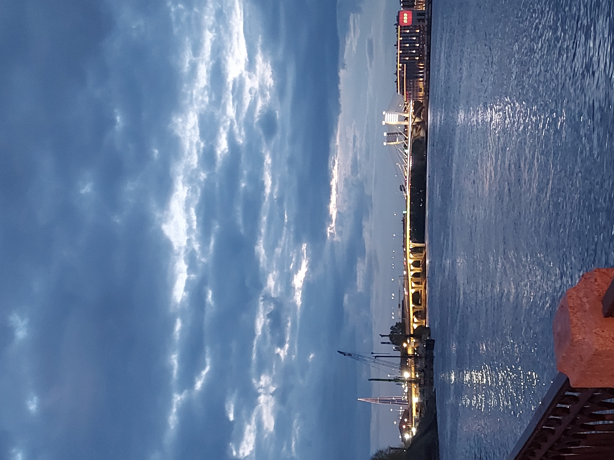The image depicts a riverfront scene in Saint Petersburg, Russia, at dusk or night. The sky is filled with dark, textured clouds. A body of water, likely the Neva River, is visible, reflecting light. A bridge with lighted arches spans the river. In the distance, a modern cable-stayed bridge with illuminated pylons is visible, along with illuminated buildings. A construction crane stands along the riverbank. A part of a stone barrier or railing is in the foreground.