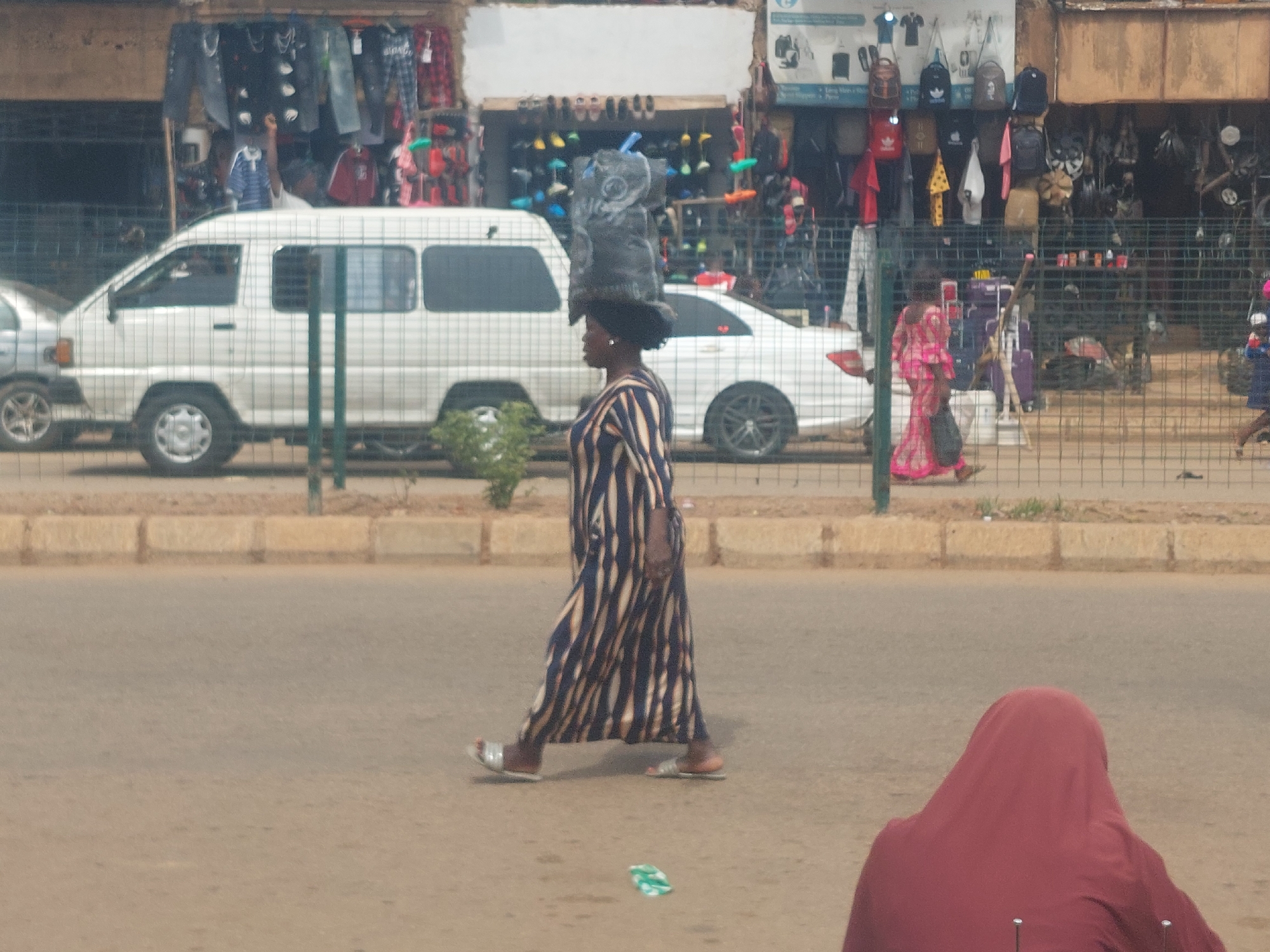 A woman wearing a long, striped dress walks on a paved road, carrying a large bag of items balanced on her head. She has dark hair and is wearing sandals. In the background, a white van and a white sedan are parked, and a shop with various goods displayed on shelves is visible. Another woman in a pink garment is walking in the distance, and a person wearing a red hijab is in the foreground, facing away from the camera. The scene appears to be in a bustling urban environment.