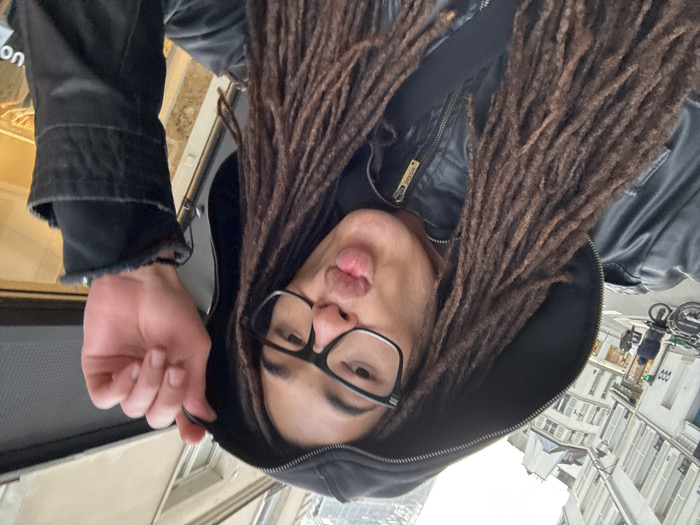 A person with long dreadlocks, wearing glasses and a black jacket with a hood, is making a kissing face. The person is outdoors. In the background, there are buildings and people on the sidewalk, possibly in a city. The photo is taken from a low angle, looking up at the person. The image is rotated and the person is upside down.