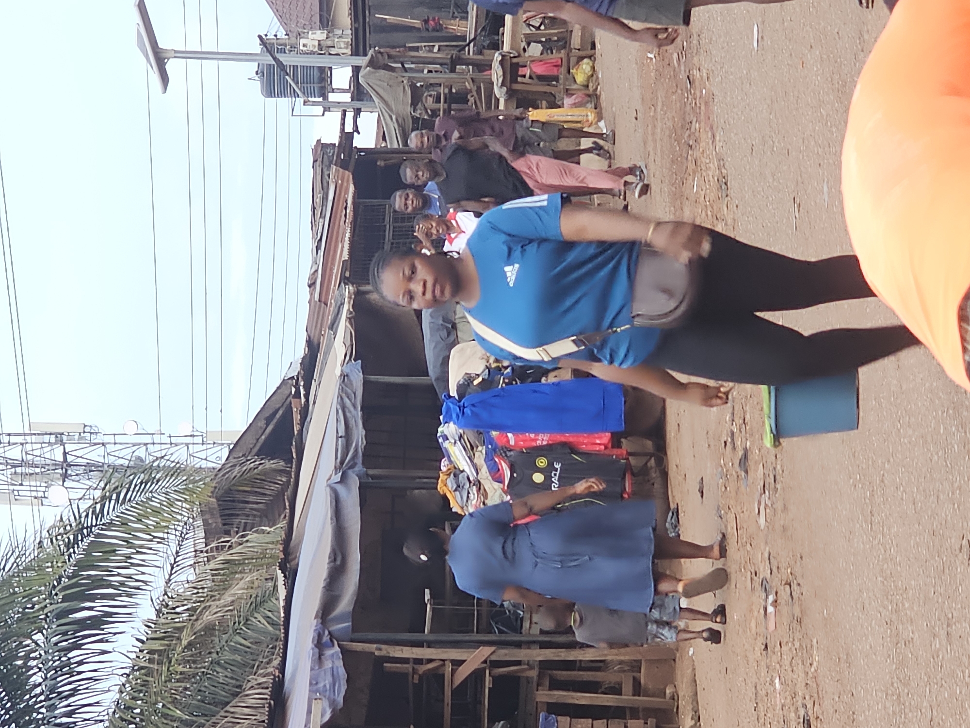 In an outdoor market in Unknown, Unknown, a woman wearing a blue t-shirt and black leggings walks towards the viewer, with a brown purse slung across her body. Behind her, a woman in a blue dress walks away from the camera, accompanied by a child. Several other individuals are visible in the background, including men in casual clothing. The market setting features makeshift stalls with corrugated metal roofs, displaying clothing and various goods. Power lines and a cellular tower are visible in the sky. The ground is unpaved and dusty.