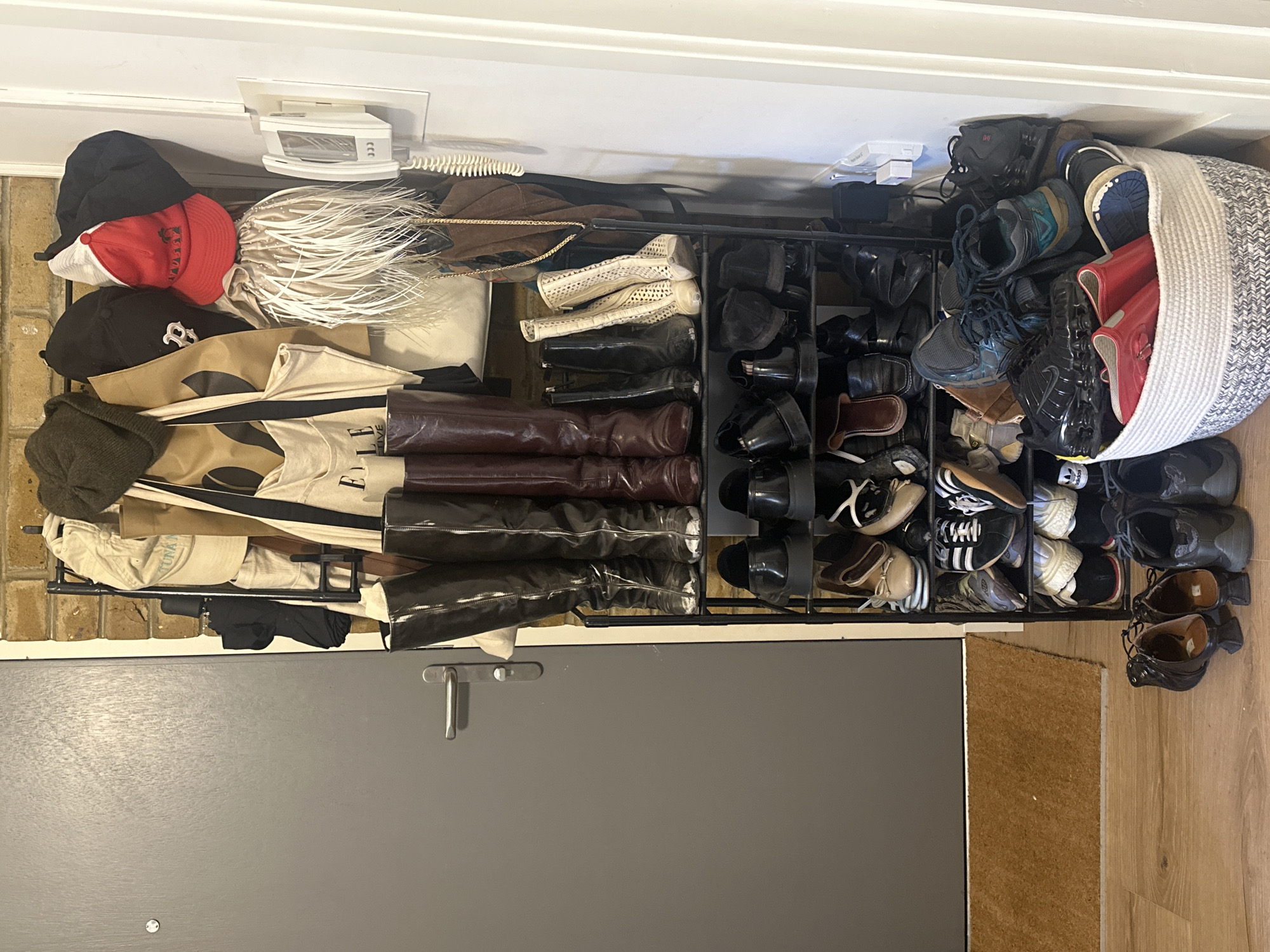 The image depicts an indoor scene featuring a cluttered entryway. A metal shoe rack is positioned against a wall, overflowing with a variety of footwear, including boots, sneakers, and dress shoes. Atop the rack are an assortment of hats, handbags, and other miscellaneous items. Adjacent to the rack is a gray door with a silver handle, and a small section of a brown doormat is visible on the wooden floor. The location is identified as London, United Kingdom.