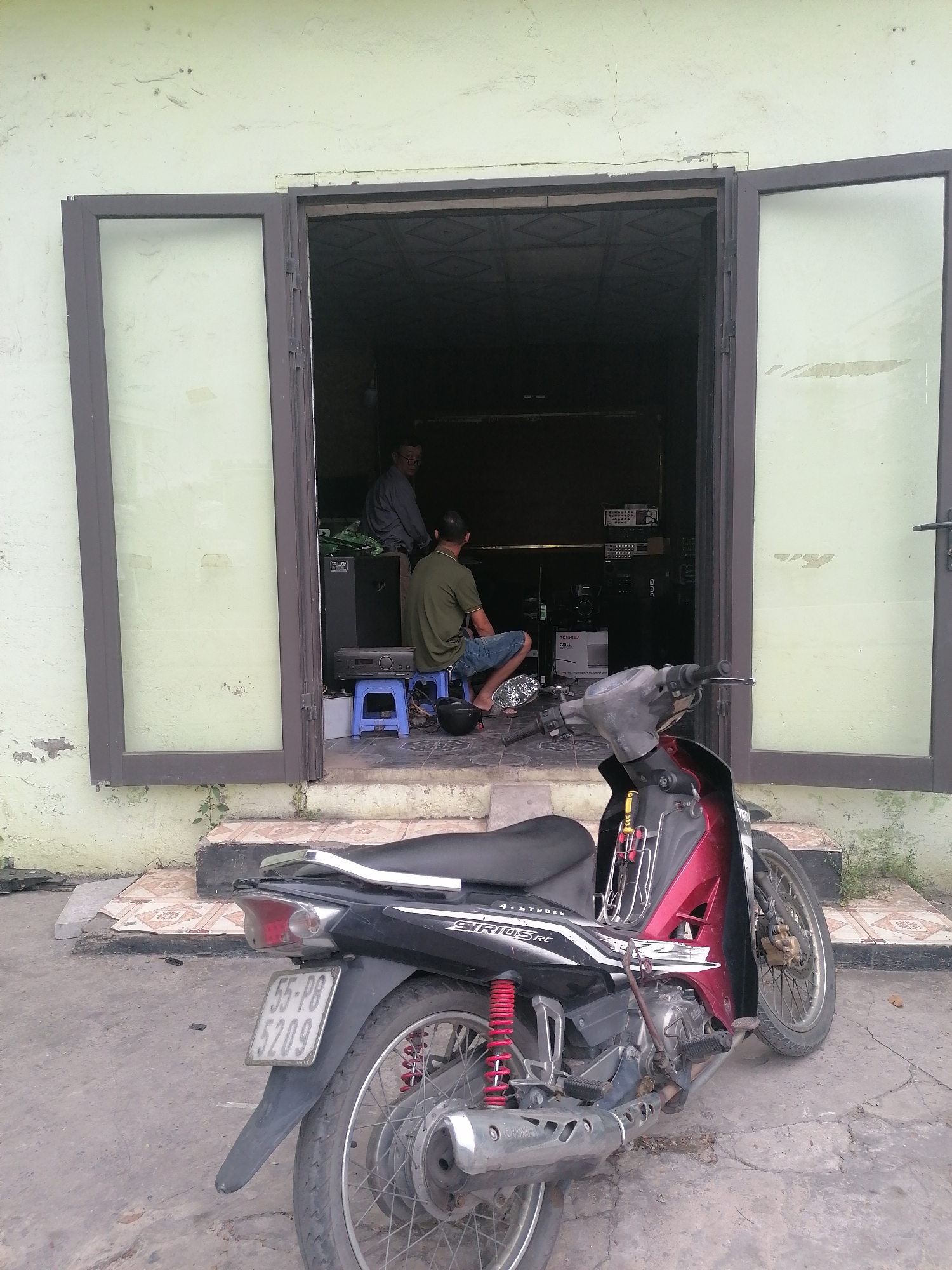 The image, taken in Ha Tu Ward, Vietnam, shows a motor scooter parked in front of an open doorway. The scooter is primarily black and red, with the license plate reading "55-P8 5209." Two men are visible inside the building, appearing to be working with audio equipment. The doorway is framed by light green walls and tiles. The environment suggests a small, possibly local business.