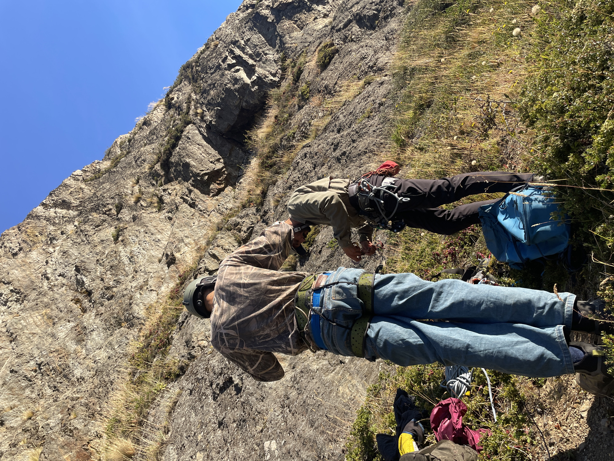 Two people are on a mountain slope near Ushuaia, Argentina. The person in the foreground is wearing a camo shirt, blue jeans, and a helmet. They have climbing gear around their waist. The second person is wearing a green jacket, dark pants, and a helmet. They also have climbing gear. A blue backpack is visible next to the second person. The mountain slope is rocky with patches of grass and vegetation. The sky is clear and blue.