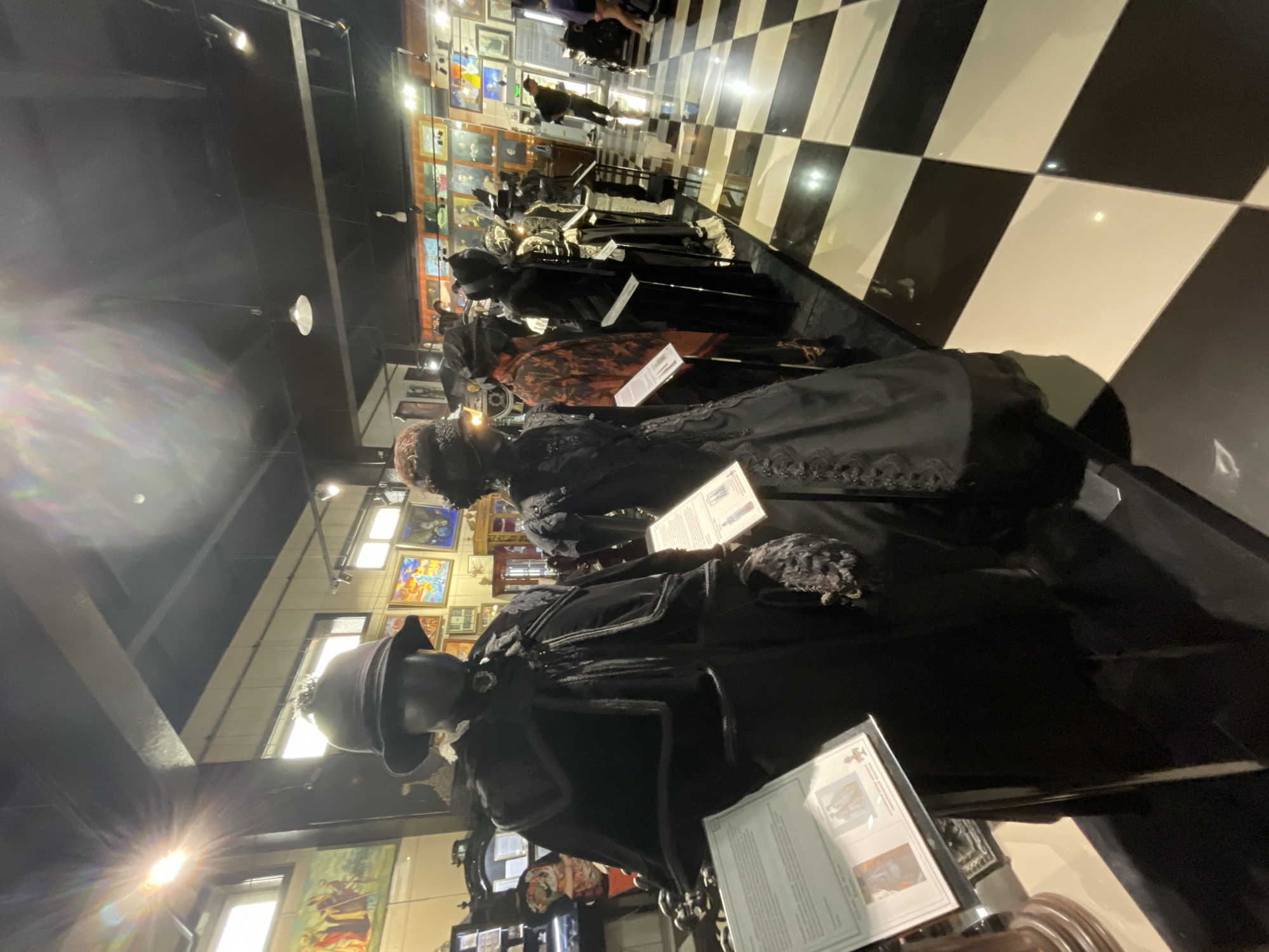 The image depicts the interior of a museum exhibition featuring mannequins dressed in historical clothing. In the foreground, a mannequin on the left wears a dark, elaborate Victorian-era outfit consisting of a coat, cape, and hat, with a descriptive placard in front of it. To its right, another mannequin is dressed in a similar dark, ornate gown. Several other mannequins in period attire are visible in a line receding into the background. The exhibition space has a black and white checkered floor. The walls are adorned with framed artwork and displays. The lighting is artificial, with visible ceiling lights and spotlights. The overall impression is of a museum showcasing historical fashion. The image is not geographically specific to Voskhod, Russia, as there are no visual cues or context provided within the image to indicate this location.