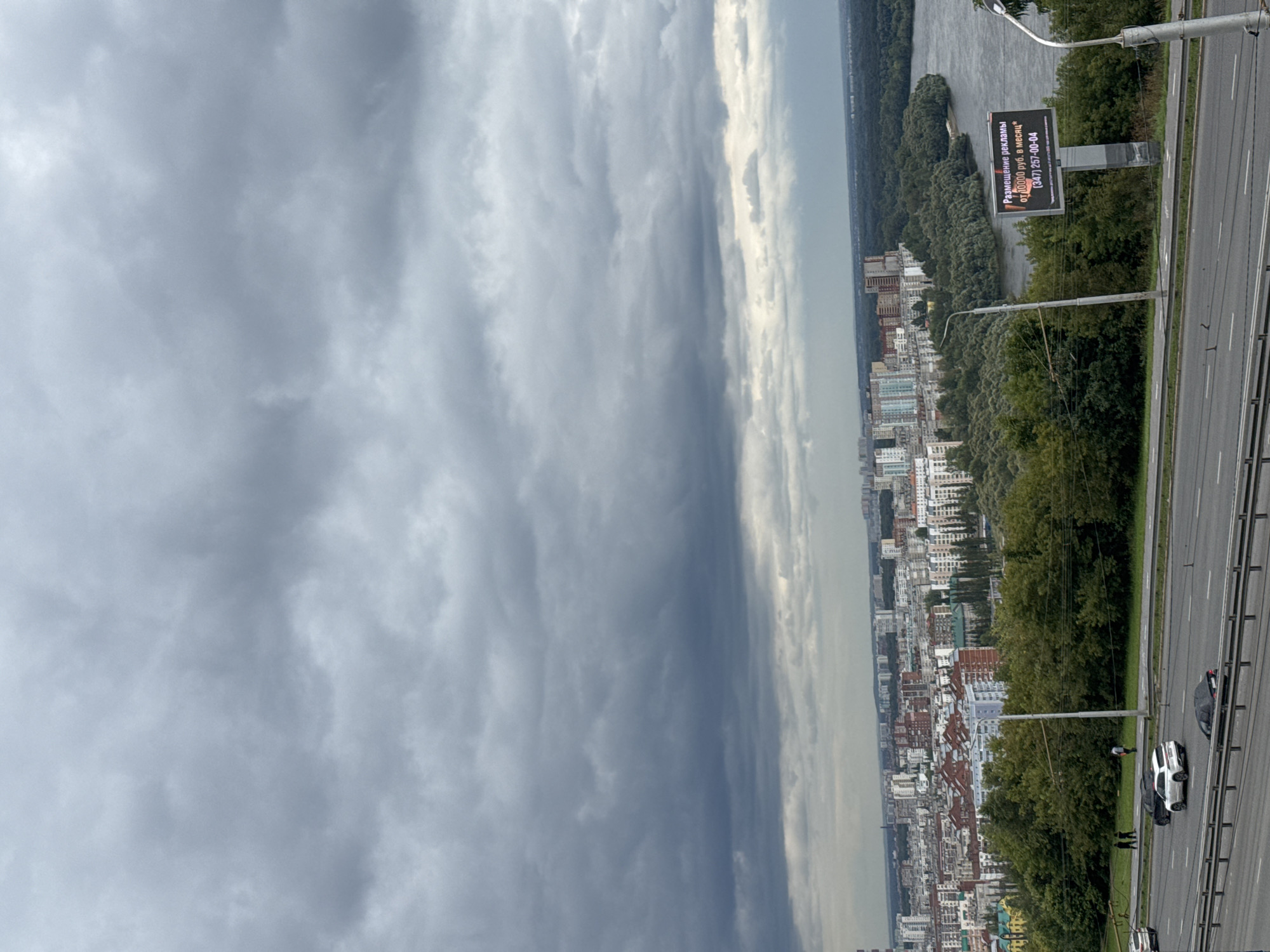 The image captures a panoramic view of the city of Ufa, Russia, with a cloudy sky dominating the upper portion. In the foreground, a multi-lane highway is visible with several cars driving on it. A white SUV and a black sedan are the most prominent vehicles. To the left of the highway, a grassy embankment is present, with a few pedestrians walking along a path. Behind the embankment, a dense green forest lines the bank of a wide river. Across the river, a cityscape unfolds, characterized by numerous apartment buildings of varying heights and architectural styles, interspersed with trees. A billboard is situated on the right side of the image, displaying an advertisement in Russian. In the distance, more buildings and trees stretch towards the horizon, under the overcast sky.