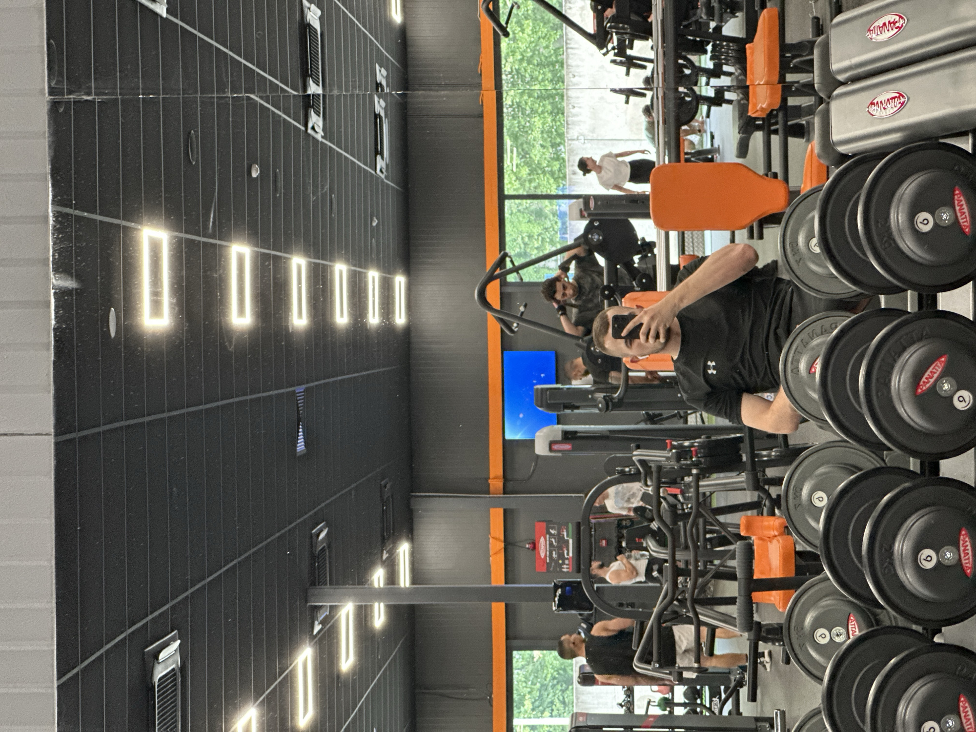 The image captures a scene inside a gym, likely located in the city of Plovdiv, Bulgaria, given the presence of people who may reside there. A man is taking a selfie using his phone, positioned in front of a rack of weights. Several people are exercising in the background, utilizing various gym equipment. The gym has a mirror spanning the length of the wall, reflecting the activity and enhancing the sense of space. The interior design features dark walls with recessed lighting. The floor appears to be tiled.