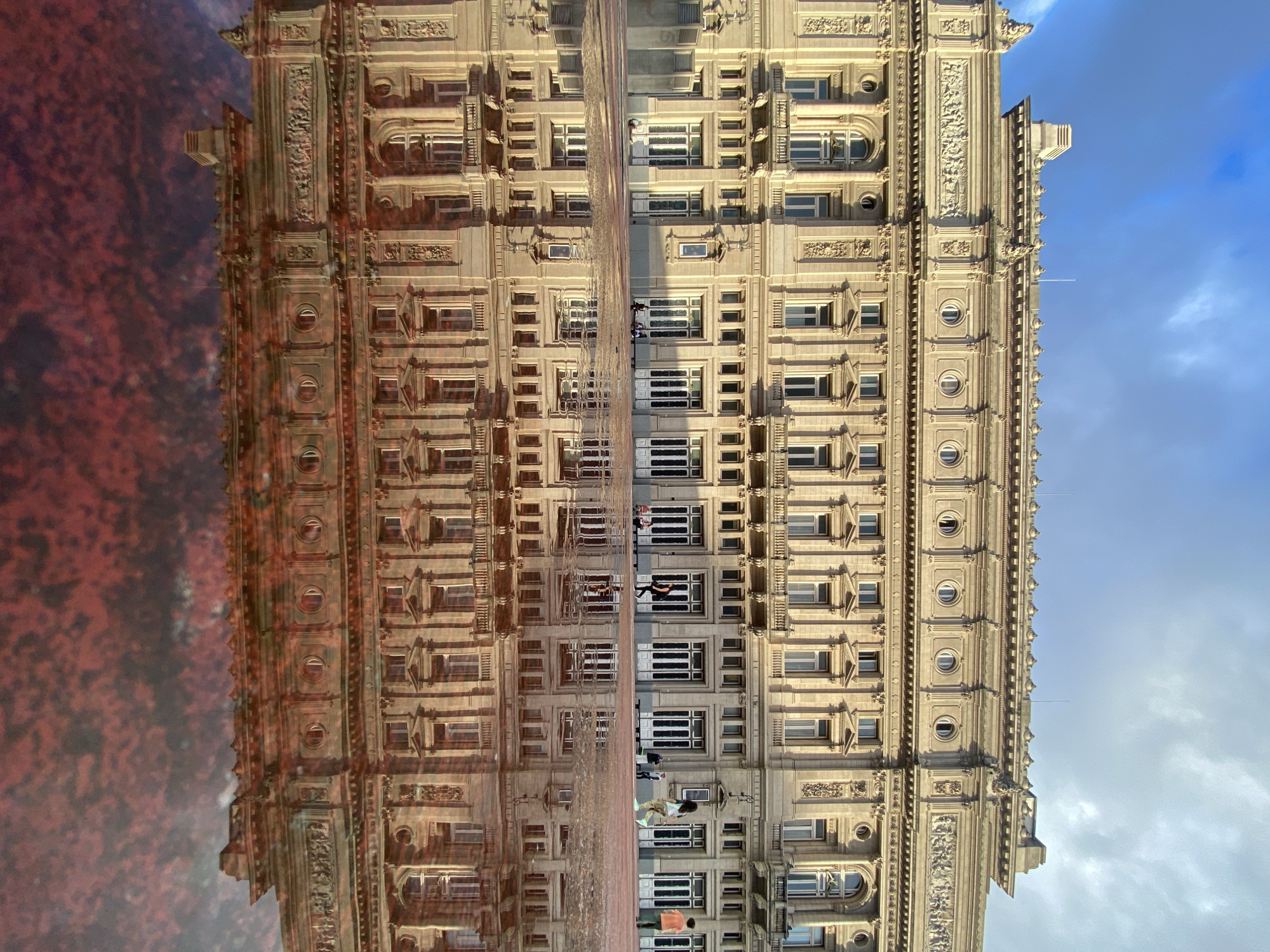 The image displays a grand, ornate building captured alongside its vibrant reflection. The building, likely situated in Buenos Aires, Argentina, appears to be made of light-colored stone or concrete, boasting a classical architectural style. It features numerous stories with a variety of window types, including rectangular, arched, and circular openings, all adorned with intricate decorative elements such as cornices, carvings, and moldings. The sky above the building is a clear blue with scattered white clouds, suggesting daylight.
On the opposite side, a prominent reflection of the building is visible in a dark, reddish, rippled surface, strongly indicating a large puddle or wet ground on a reddish pavement. This reflection is somewhat distorted by the water's movement. Small figures of people can be seen on what would be the ground level in front of the building, as well as their reflections. They appear to be walking or standing, adding a sense of scale and life to the urban scene. The perspective is unusual, with the building seemingly laid on its side, creating a striking visual symmetry with its reflection. No specific text is discernible on the building or in the scene.