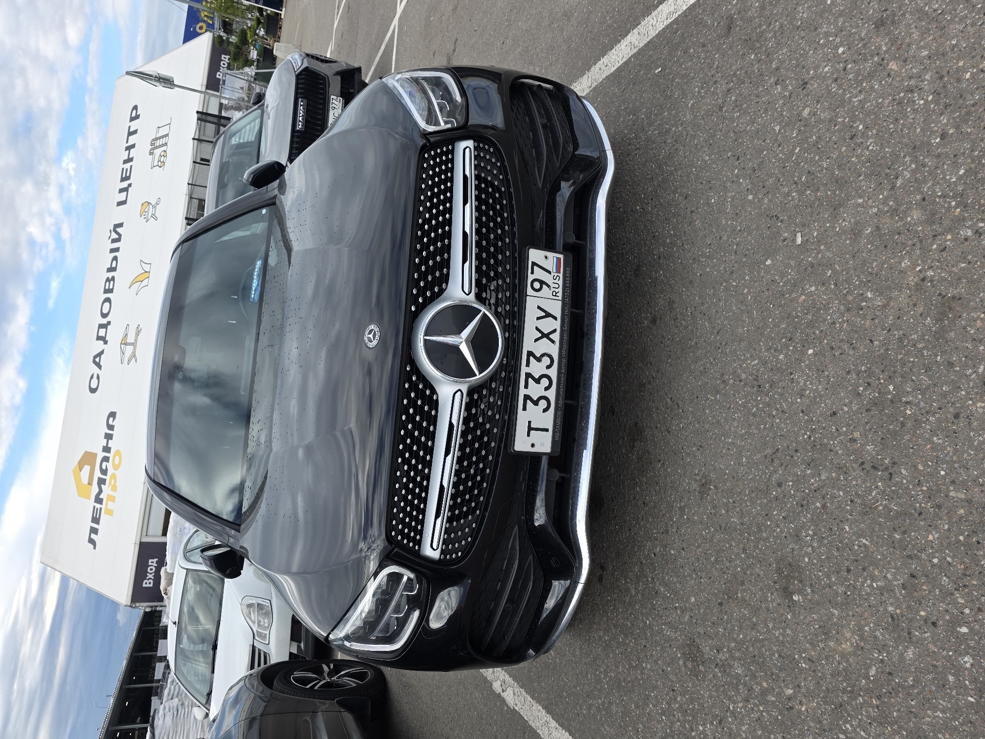This media file captures a close-up, low-angle view of the front of a black Mercedes-Benz SUV parked outdoors. The vehicle's grille, headlights, and license plate are prominently featured. The license plate reads "T333XY 97" with "RUS" below it. The background shows a white building with Cyrillic text, including "САДОВЫЙ ЦЕНТР" (Garden Center) and "Вход" (Entrance). A yellow logo with a house and the word "ЛЕМАНА" is also visible on the building. Another car, possibly a white sedan, is partially visible behind the Mercedes. The ground is asphalt, marked with white lines. The sky appears overcast with clouds. The overall impression is of a daytime scene, likely in a parking lot of a commercial establishment. No people are visible in the frame.