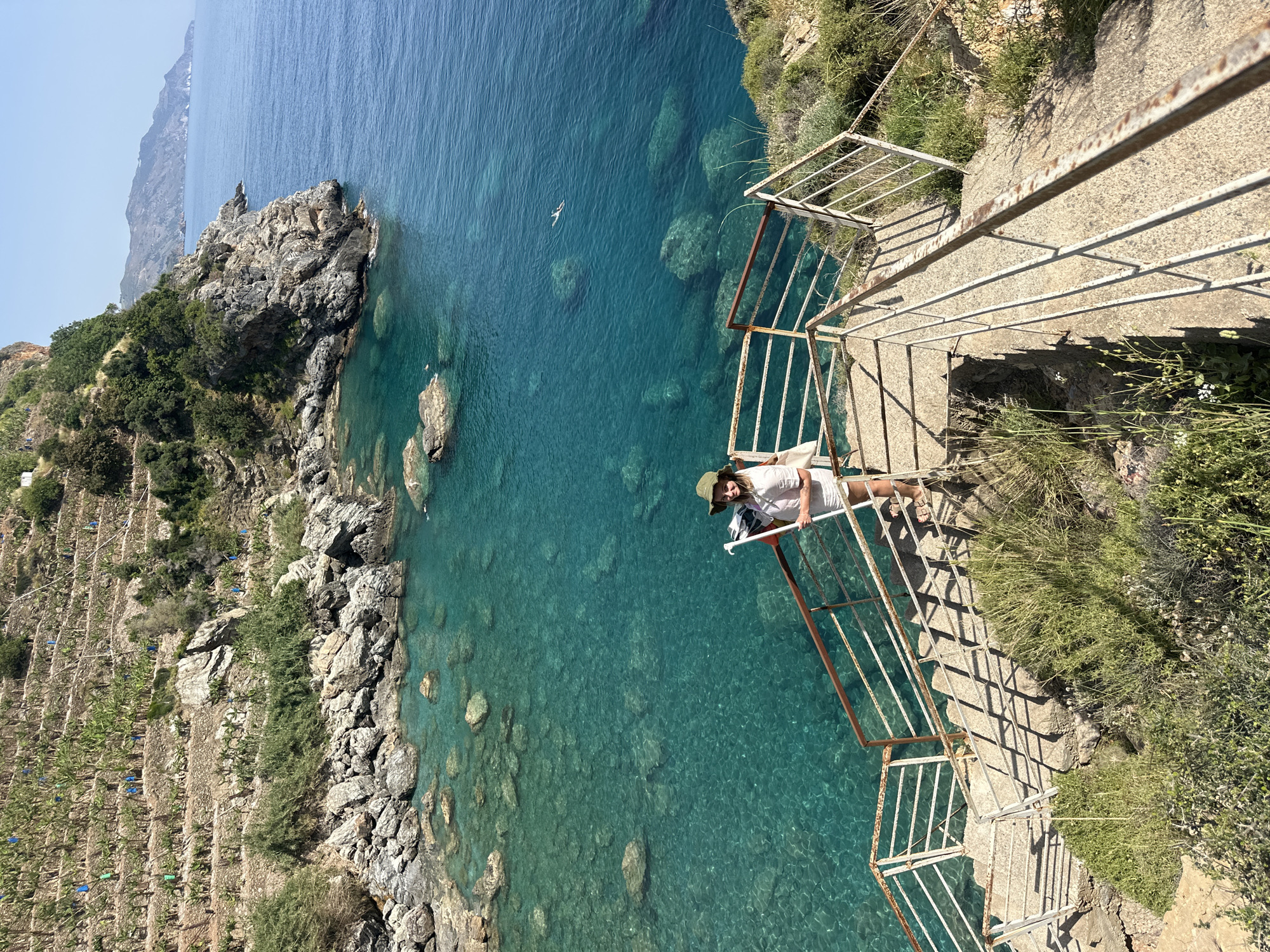 A medium, eye-level shot shows a woman wearing a white outfit and a hat, sitting on a railing overlooking the Mediterranean Sea at Gazipaşa, Turkey. The camera is positioned above the woman, showing the scene from a high angle. The woman leans forward slightly, looking towards the camera. The railing is white with some rust, and it borders a set of stone steps leading down the cliff. The sea is a deep turquoise, and visible rocks and the seabed are seen through the clear water. The coast to the left is rocky and covered with green vegetation, including agricultural terraces. In the distance, part of the coastline extends into the sea. The sky is light blue and clear.
