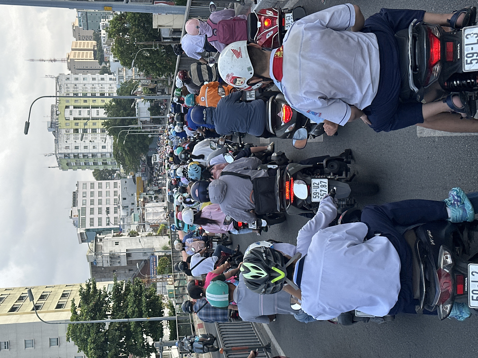 The image captures a congested street in Ho Chi Minh City, Vietnam, filled with numerous motorcycles. The vehicles are tightly packed together, indicating heavy traffic, with many riders wearing helmets. Streetlights line the road, and buildings of varying heights form the cityscape in the background. The sky is overcast. The closest vehicles are being operated by young people in school uniforms. License plates on some of the vehicles are visible, displaying the alphanumeric pattern common in Vietnamese vehicle registration.