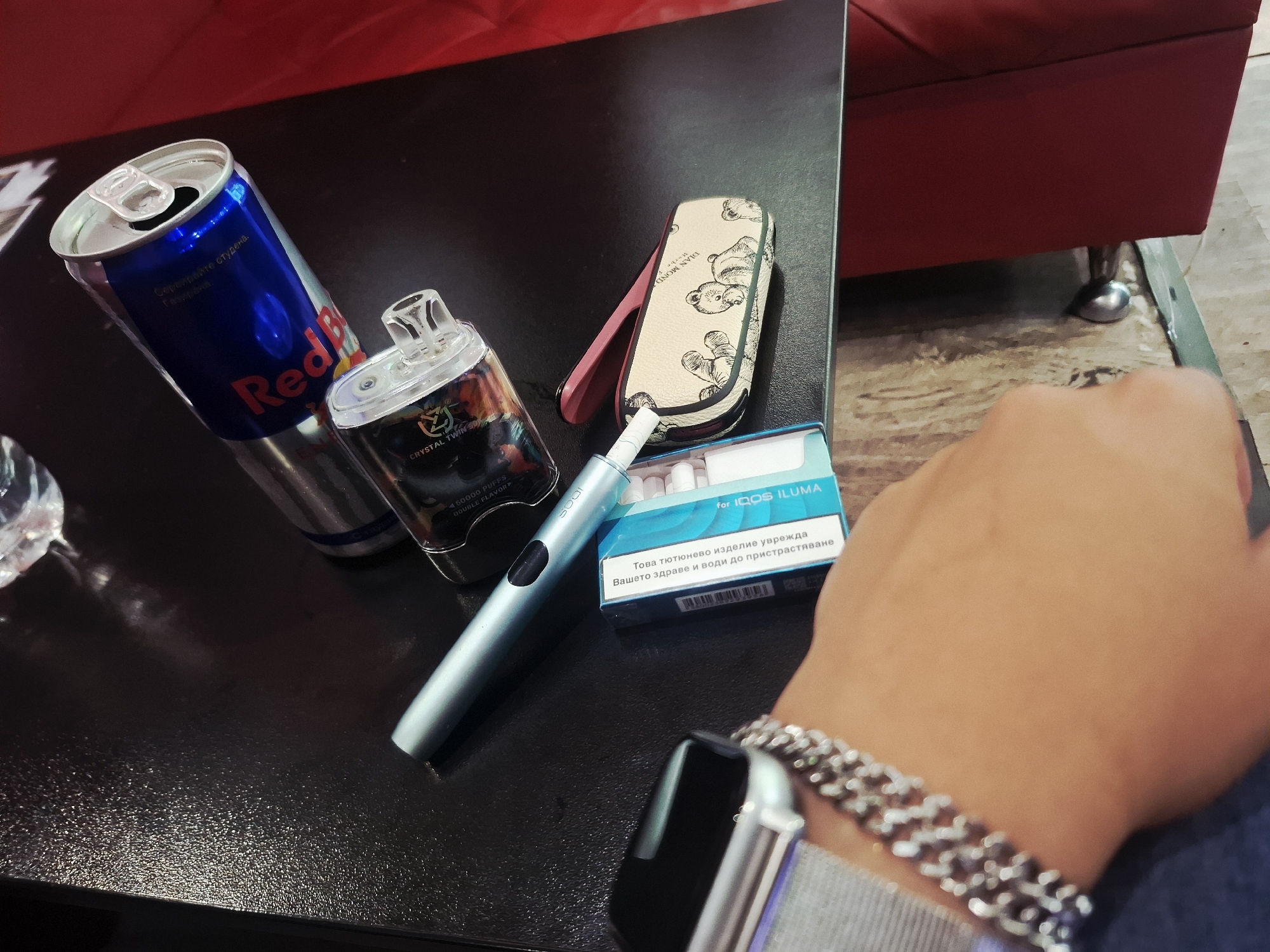 The image presents an indoor scene, likely a casual lounge or private setting, featuring several personal items arranged on a dark, textured table. In the foreground, a person's right hand, adorned with a silver chain bracelet and a silver smartwatch, rests partially visible.
Dominating the left side of the table is an opened blue and silver Red Bull energy drink can, with Cyrillic text "Сервирайте студена. Газирана." (Serve cold. Carbonated.) visible on its label. Adjacent to the Red Bull is a clear, rectangular vape device, featuring the branding "CRYSTAL TWIN S50000 PUFFS DOUBLE FLAVOR." A sleek, light blue or silver IQOS ILUMA device lies diagonally across the table, its name "ILUMA" faintly visible. Alongside it, a light blue and white box of tobacco sticks for IQOS ILUMA is prominently displayed, bearing the health warning in Bulgarian: "Това тютюнево изделие уврежда Вашето здраве и води до пристрастяване" (This tobacco product harms your health and leads to addiction). Behind these smoking accessories, a cream-colored IQOS case with a pink hinge and black teddy bear illustrations is partially open, with "ELIAN MOND" text visible.
In the background, a section of a red upholstered chair or sofa is visible, suggesting a comfortable environment. The floor beneath the furniture appears to be a light-toned, possibly tiled or stone surface. The presence of an opened energy drink and electronic smoking devices indicates a moment of leisure or a break. The prominent Bulgarian text on both the Red Bull can and the IQOS product packaging serves as a strong cultural and linguistic cue, aligning with the given location context of Mezdra, Bulgaria.