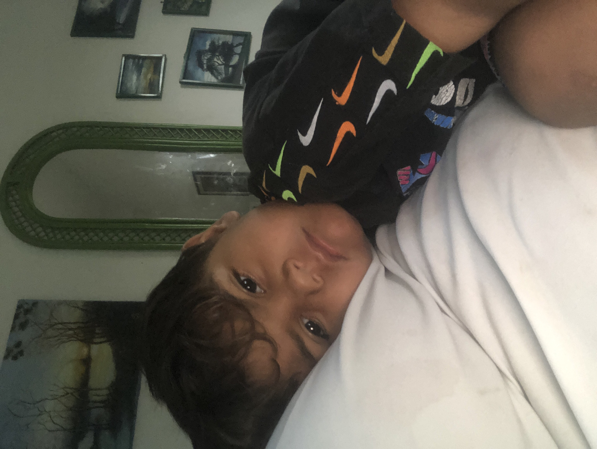 A child with dark hair and brown eyes is lying on a white surface, possibly a bed, with their head tilted. The child is wearing a black hoodie with colorful Nike swoosh logos. The background shows a wall with several framed pictures and a large, ornate green mirror. One of the pictures appears to be a landscape painting, possibly of trees. Another framed artwork is a smaller, abstract piece. The overall lighting suggests an indoor setting, possibly during the day. The child's expression is neutral, and the image is taken from a close-up perspective, focusing on the child's face and upper body. The orientation of the image is portrait, but the content is rotated.