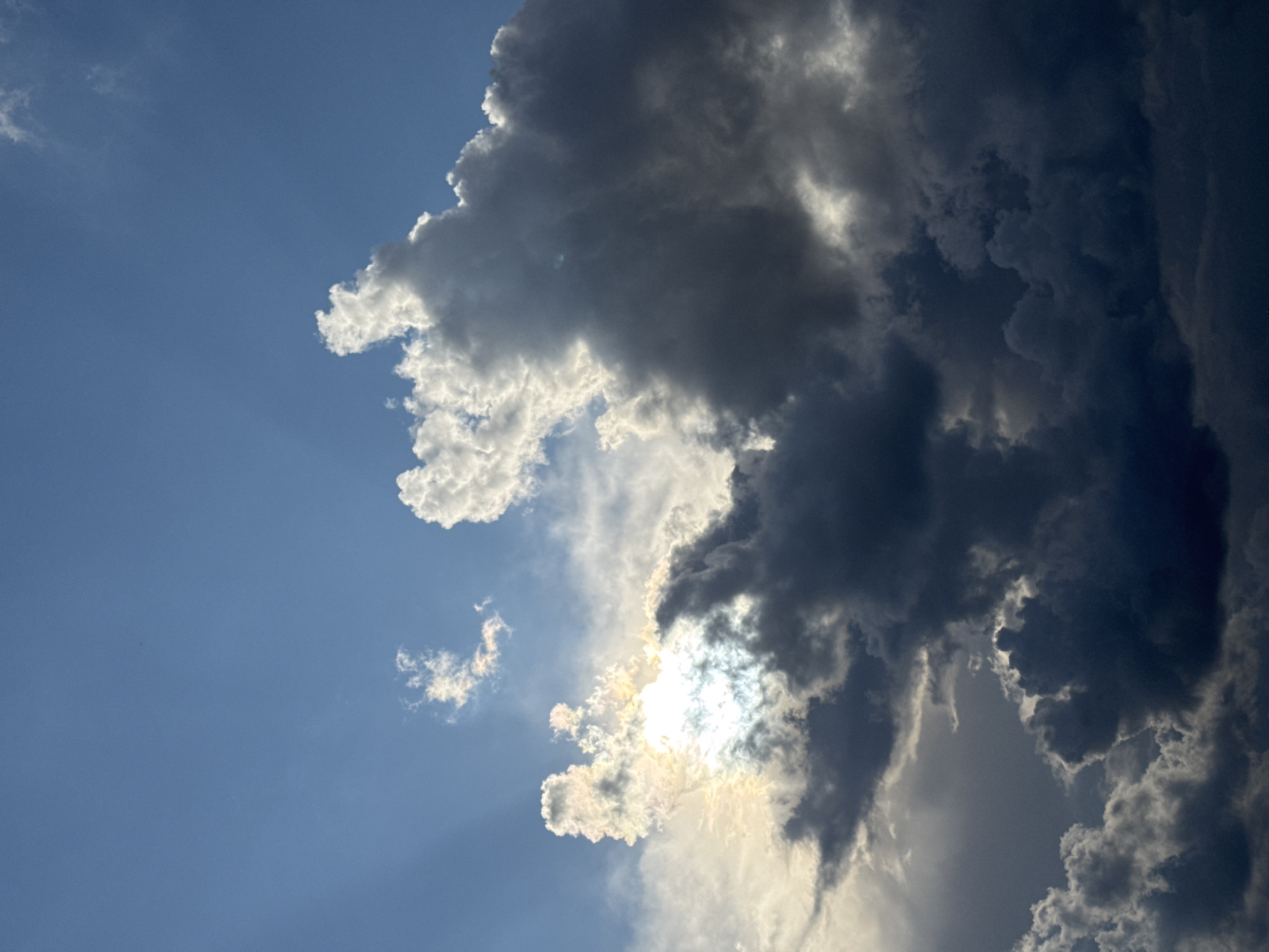 The image shows a dramatic sky filled with a mix of dark, heavy storm clouds and bright, sunlit clouds. The sun is partially obscured by the clouds, creating rays of light that pierce through. The sky transitions from a clear blue at the top to darker shades of grey and black as it descends. The clouds are voluminous and textured, with sharp contrasts between light and shadow. There are no people, buildings, or any identifiable landmarks present in the image. The scene is solely focused on the atmospheric conditions of the sky. The image does not contain any visual information that can be used to determine its location in the city of Minsk, Belarus.