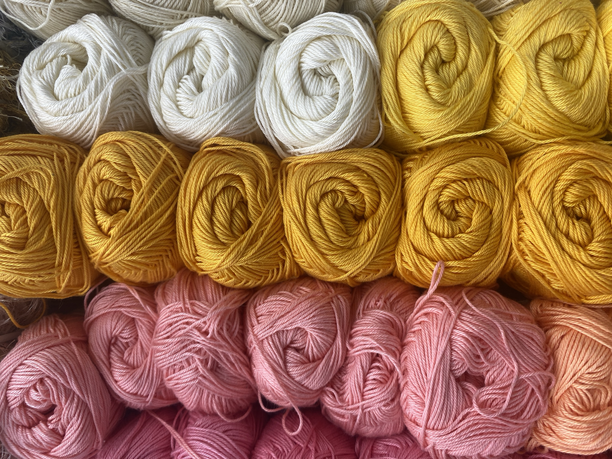 The image is a close-up, overhead shot of many tightly wound balls of yarn arranged in rows. The yarn is primarily in three colors: pink, yellow, and white, with some slight variations within each color. The balls are stacked horizontally and vertically, creating a textured grid. No people, actions, or interactions are visible. The environment appears to be a retail setting, likely a craft or yarn store, but its specific location in Krasnodar, Russia, cannot be determined from the image alone.