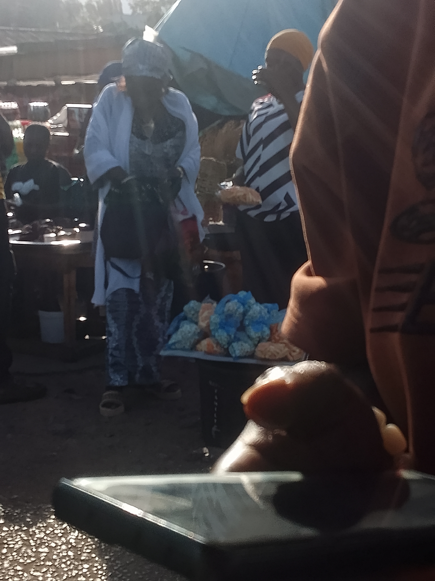 The image, taken in Bukuru, Nigeria, shows a street scene likely at a market. In the midground, a woman in a white overcoat, patterned dress, and head covering stands near a table with bags of blue-wrapped items, possibly food. Another woman with a yellow head covering and striped shirt stands to the right, holding something in her hand. Other people are visible in the background. In the foreground, a mobile phone partially obstructs the view. A blurred arm is near the phone. The ground appears to be a rough, uneven surface. The overall lighting indicates the scene is outdoors.