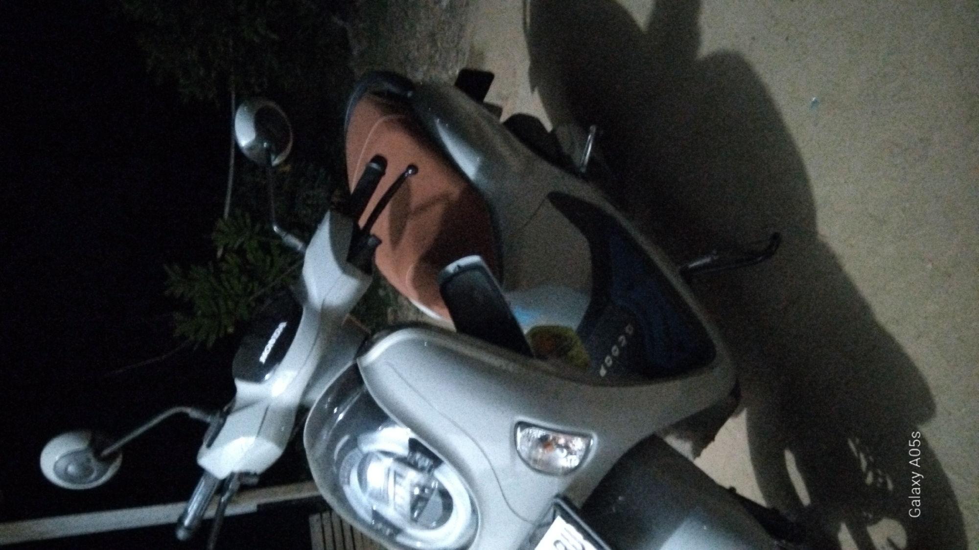A silver Honda scooter is parked outdoors at night in Rambongpayong, Indonesia. The scooter is facing left and is partially illuminated by what appears to be street lighting or ambient light. The scooter has a brown seat and a prominent headlight assembly. The background is dark, with the shapes of trees and foliage visible, indicating an outdoor environment. The ground beneath the scooter is a dirt or gravel surface. A shadow of the scooter is cast on the ground. The "Galaxy A05s" watermark is visible in the bottom left corner of the image.