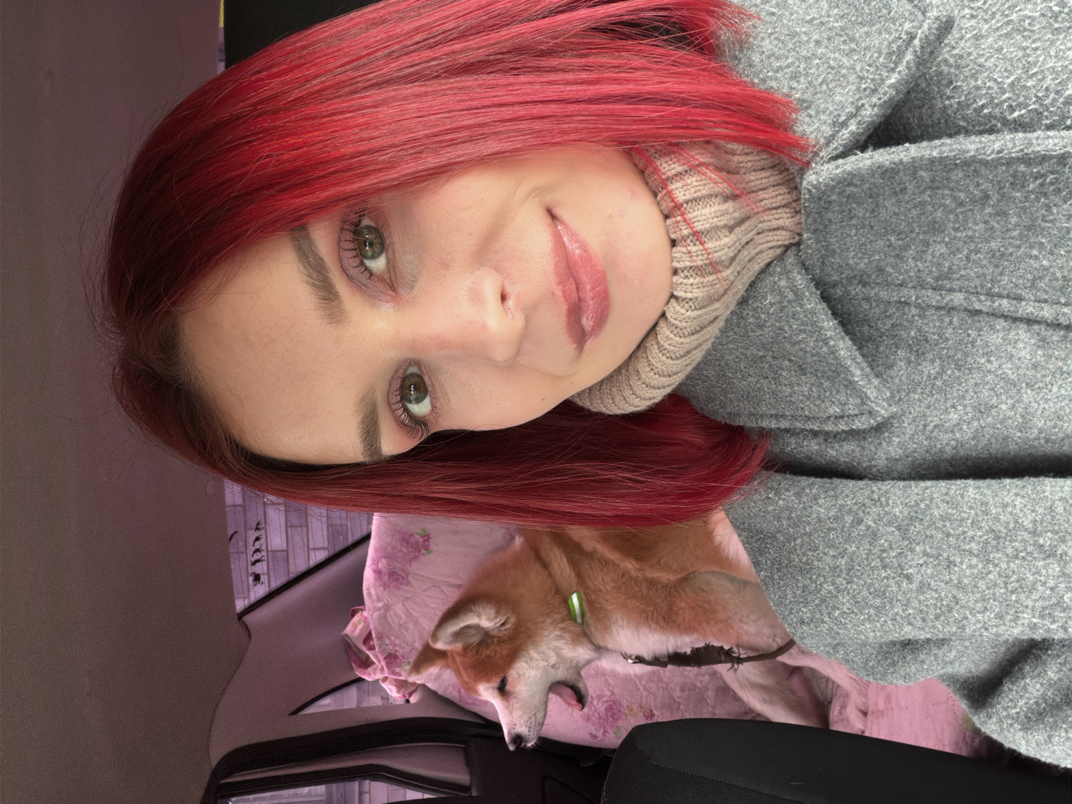 A young woman with vibrant, long red hair and green eyes is captured in a selfie taken inside a car. The image is rotated 90 degrees clockwise. She smiles gently at the camera, wearing a light gray wool coat over a beige ribbed high-neck sweater, suggesting cool weather. Her makeup includes visible eyeshadow, mascara, and lip gloss.
Beside her, a small to medium-sized dog, with light brown and cream fur and a light green collar, lies relaxed on what appears to be a car seat covered with a pink floral-patterned blanket. The dog's tongue is slightly out, indicating it is resting or possibly napping.
The background shows elements of a car interior, including a dark headrest and seat upholstery. The lighting is soft and natural, typical of daytime. The scene conveys a calm and affectionate moment between the woman and her pet, set within a car in Chaikovsky, Russia. No visible text is present in the image.