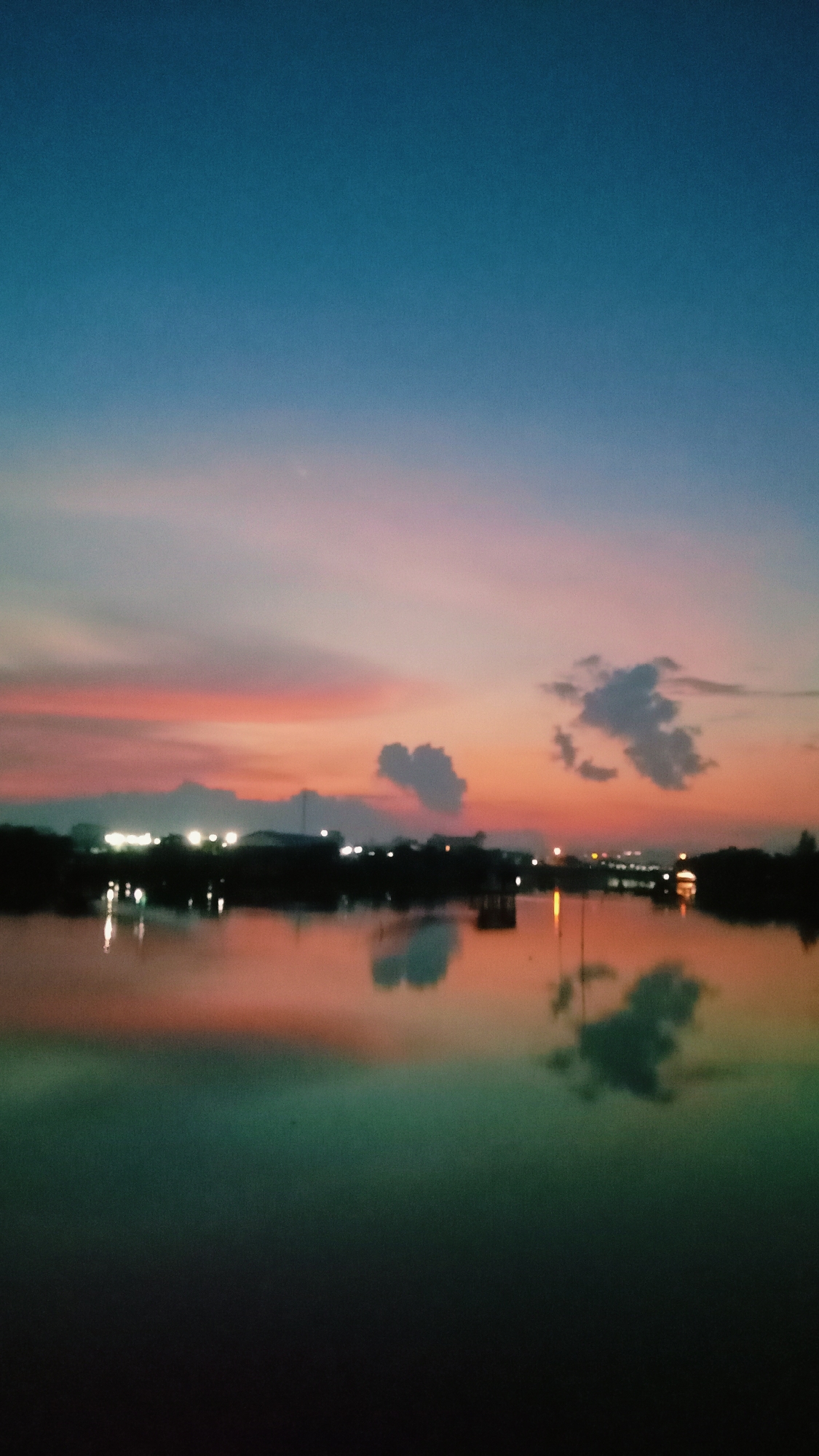 This image captures a serene dusk or early night scene over a large body of water, likely a lake or wide river, in Lhokseumawe, Indonesia. The sky displays a striking color gradient, transitioning from a deep blue at the top to vivid shades of orange, pink, and red near the horizon, indicative of a sunset. Scattered, dark clouds are visible against the colorful sky, casting distinct reflections on the water's surface. The water mirrors these hues, appearing orange-pink near the distant shore and gradually deepening into a greenish-blue in the foreground.
Along the distant horizon, a silhouette of buildings or structures lines the water's edge, punctuated by numerous artificial lights that twinkle across the landscape and reflect onto the water. A few indistinct structures or objects, possibly small boats or jetties, are faintly visible on the water, with their dark forms and reflections breaking the surface. The overall impression is one of tranquil beauty at the transition from day to night, with no people or overt activities visible. The sky is clear with some clouds, suggesting calm weather. There is no visible text in the image. The image has a slightly soft focus, adding to its atmospheric quality.