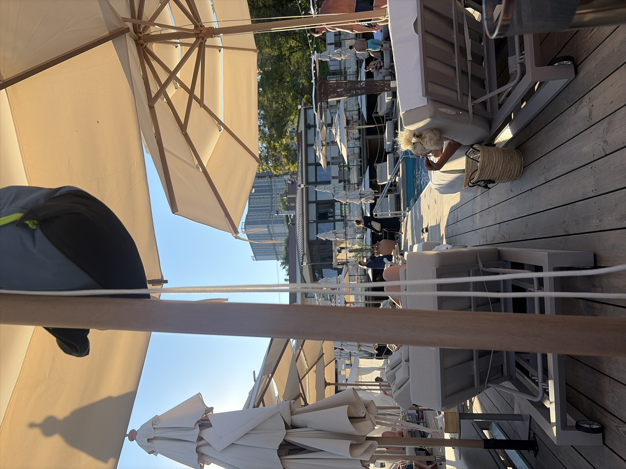 This is a wide-angle, eye-level shot of a sun-drenched outdoor lounge area, likely a beach club or resort, possibly in Odesa, Ukraine given its coastal setting and architecture. The scene is dominated by beige canvas umbrellas and lounge chairs set on a weathered wooden deck. In the foreground, a large wooden pole supporting an umbrella is prominently featured.
Several individuals are visible in the mid-ground. On the left, multiple folded umbrellas and empty lounge chairs suggest a relaxed atmosphere. In the center, a person in dark clothing stands near a swimming pool, seemingly engaged in conversation. To the right, a man with a muscular physique, wearing dark swim trunks, stands near the pool. Further back, other guests are seated on lounge chairs or walking around. A woman with blonde, curly hair is reclined on a lounge chair in the right foreground, holding a phone to her ear. A woven straw bag sits beside her chair.
The background reveals more of the lounge area, including more umbrellas and lounge chairs, leading to a low-rise building with a tiled roof and some modern high-rise buildings in the distance, suggesting an urban coastal environment. Trees line the edge of the property. The lighting indicates a sunny day, with long shadows cast across the wooden deck. The water in the pool is visible and appears clear. The overall impression is one of leisure and relaxation in a resort-like setting.
