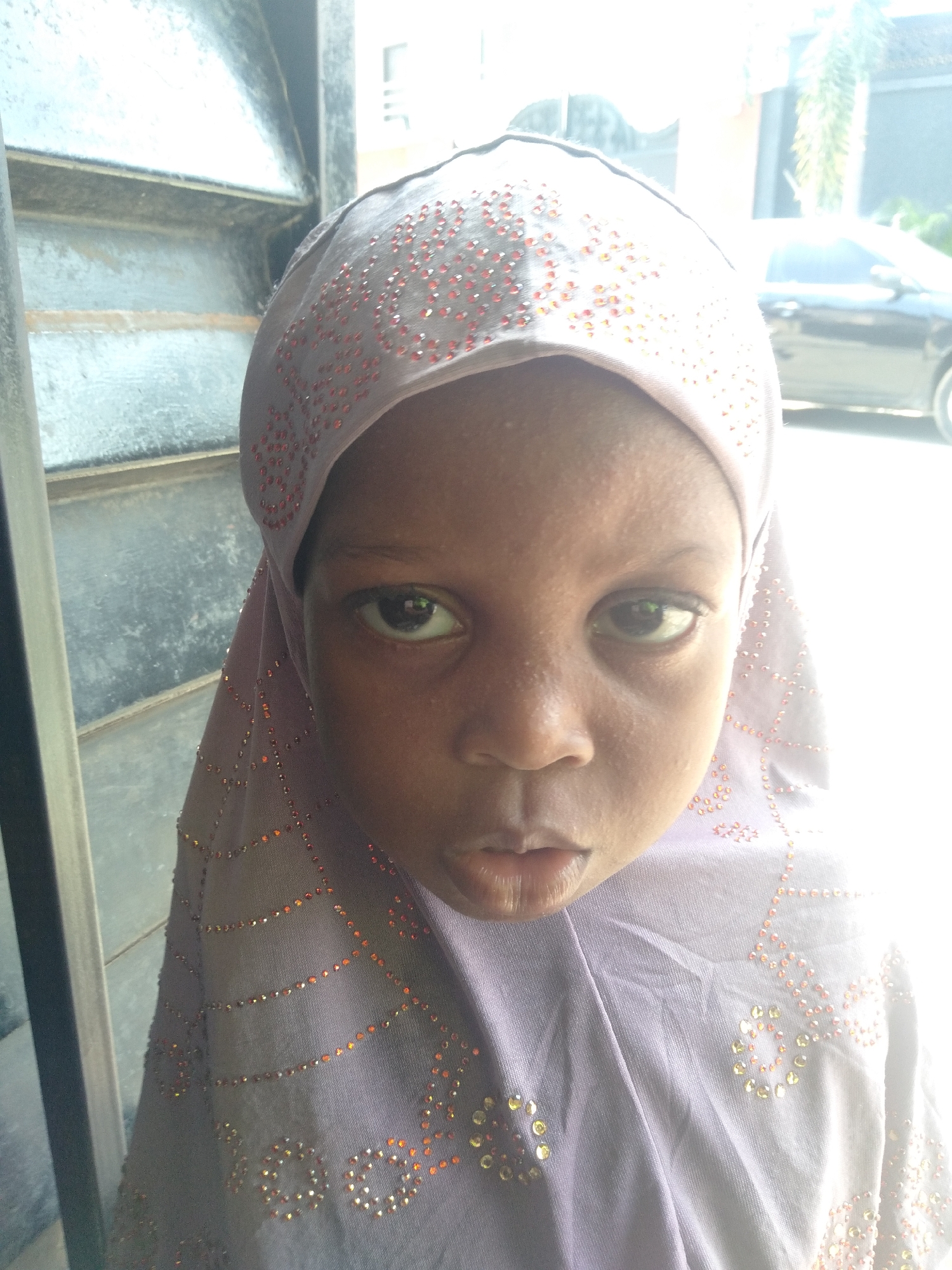 A close-up shot depicts a young child wearing a lavender headscarf with orange and silver sequins. The child faces forward with a neutral expression. To the left, a partially visible structure with vertical lines suggests a building or window. Behind the child, a black car is seen, along with greenery, indicating an outdoor setting. The location is in Shangisha, Nigeria.