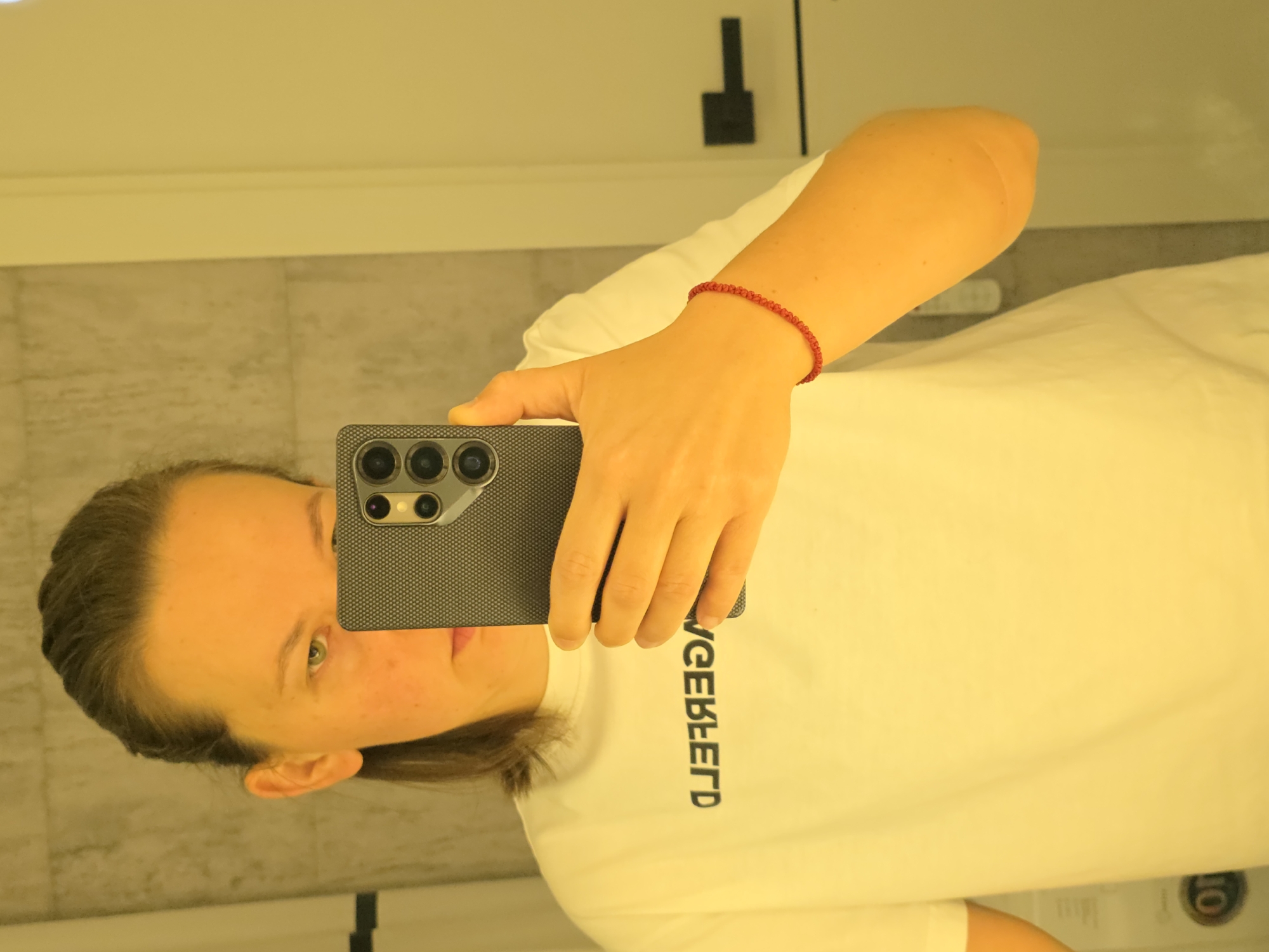 A person is taking a mirror selfie in a room that appears to be a bathroom. The person is wearing a white t-shirt with black text that is backward and appears to read "WICKEBEERD". They are holding a black smartphone with a multi-lens camera to take the photo. The person has their hair pulled back and is wearing a thin red string bracelet on their right wrist. The background shows a tiled wall and a yellow-lit door with a black handle. This image was taken in Rostov-on-Don, Russia.