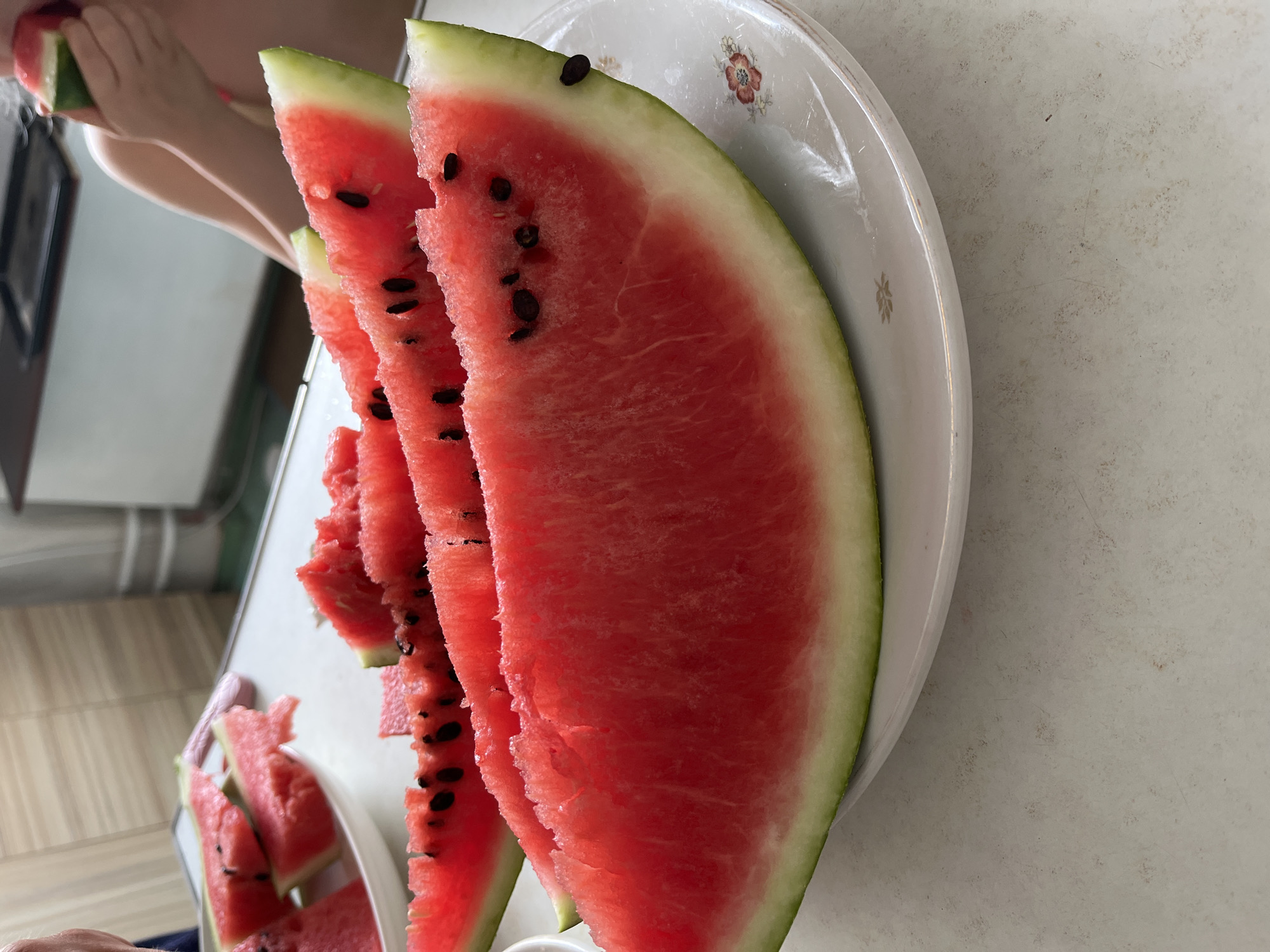 The image depicts slices of watermelon arranged on a white plate on a light-colored table. In the background, a person is partially visible, holding a small piece of watermelon. The scene appears to be indoors, possibly a kitchen or dining area, with a countertop and cabinets visible in the distance. The lighting suggests a bright, natural light source. The image is taken from a close-up perspective, focusing on the watermelon. The location information provided, Komsomolskoye, Ukraine, is external to the visual content of the image itself.
