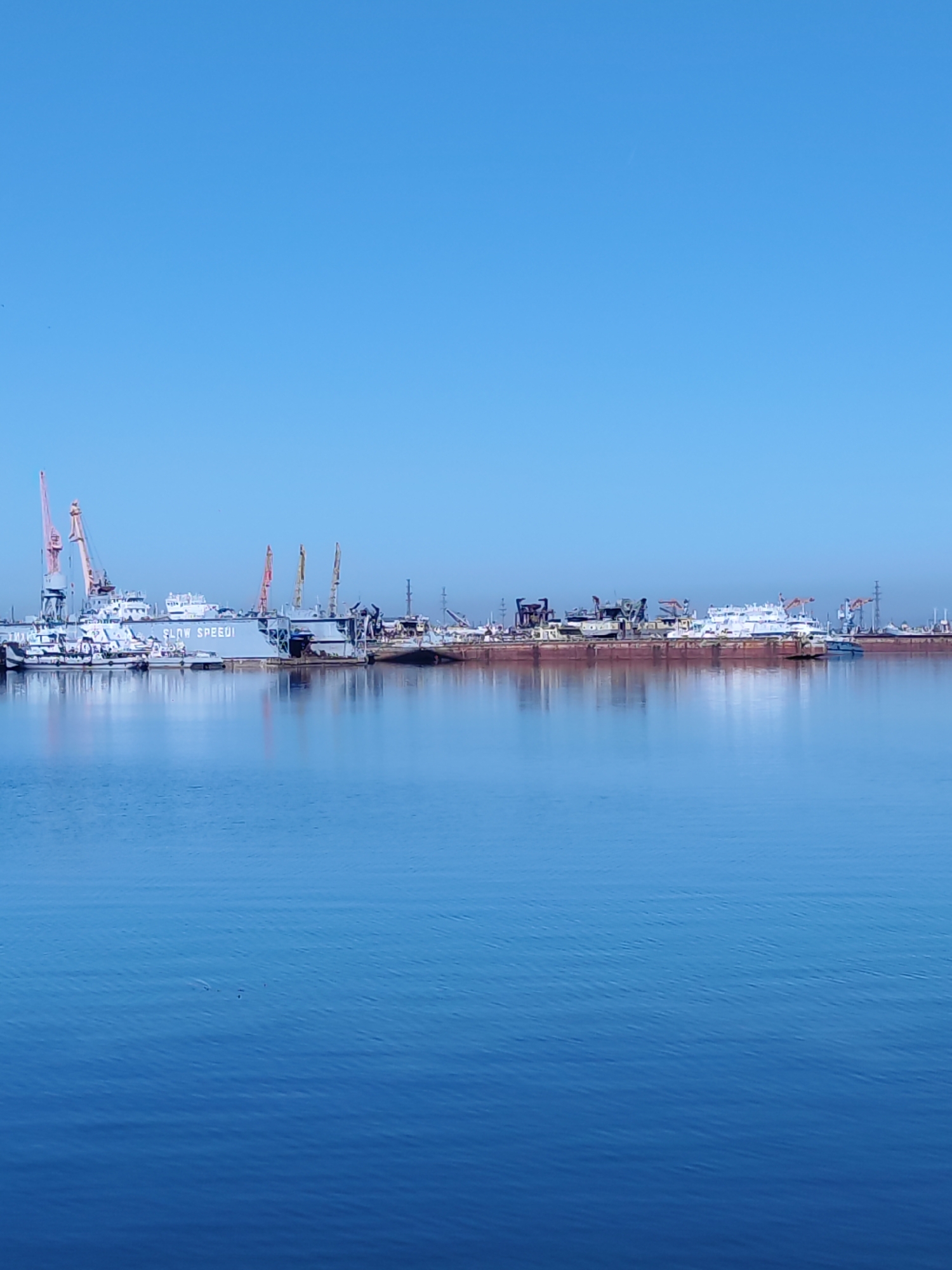 The image depicts a harbor or shipyard scene in the city of Unknown, Unknown. A body of blue water dominates the foreground. In the midground, various ships and structures are visible, including what appears to be cranes and industrial equipment. The background is a clear, light blue sky. The vessels vary in size and configuration. The overall scene appears to be a busy industrial area.