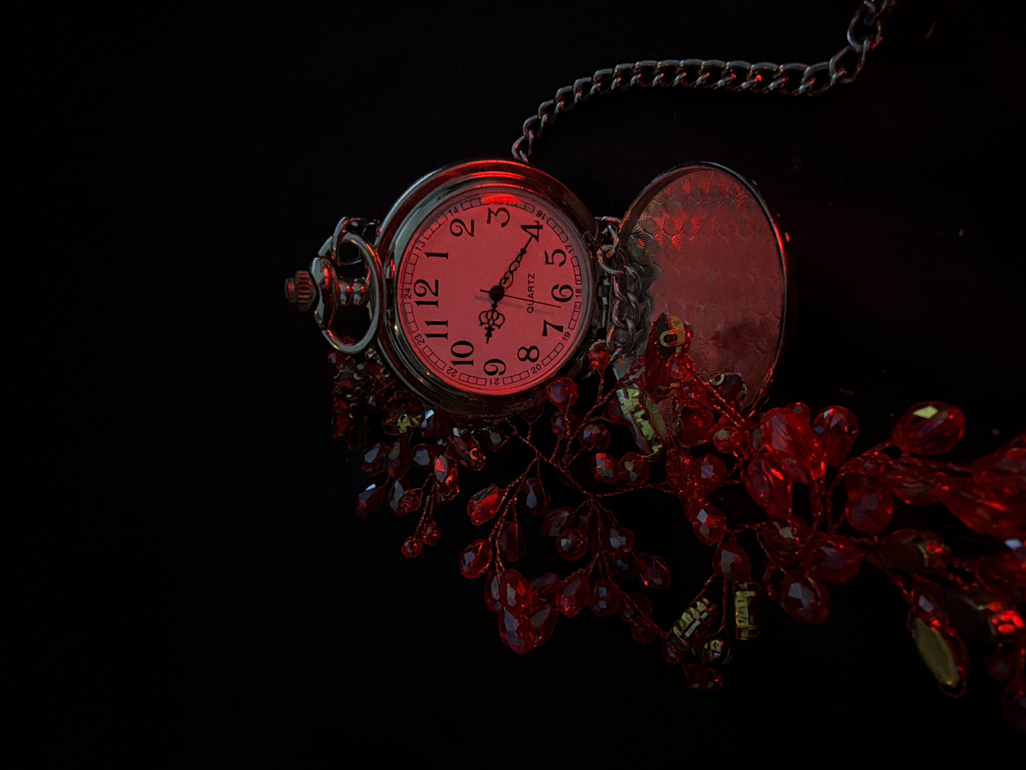A pocket watch with a red face and silver chain lies open, next to a cluster of red beaded jewelry, all on a black background. The watch face is illuminated by a red light, casting a crimson hue on the surrounding objects. The time on the watch is approximately 9:45. The jewelry appears to be made of faceted beads, some teardrop-shaped and others more rounded. The scene is lit with dramatic, low lighting, creating a stark contrast between the illuminated objects and the dark background. There are no people present in the image.