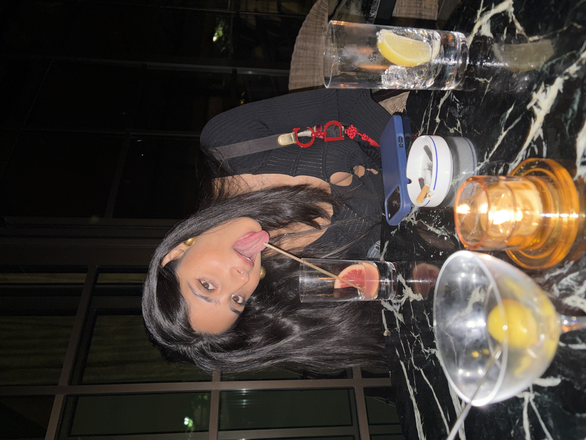A young woman with long, dark hair is seated at a black marble table, sticking her tongue out with a wooden skewer. She is wearing a black, ribbed top with cut-outs. In front of her are two glasses, one containing a pink fruit slice and ice, and another with a lemon slice and ice. A cocktail glass with a yellow olive and a spoon is also on the table, along with an ashtray containing cigarette butts and a lit candle in an orange holder. A blue smartphone is placed near the ashtray. The background is dark with large windows showing faint lights, suggesting an indoor setting at night.