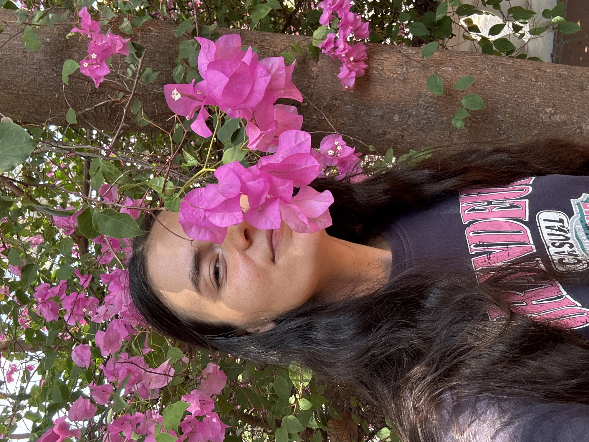 A woman is standing in front of a bougainvillea tree in what is possibly Mui Ne, Vietnam. She is facing the camera with the bougainvillea flowers partially obscuring her face. She has long, dark hair and is wearing a dark T-shirt with a pink and white design. The bougainvillea is in full bloom with bright pink flowers. The trunk of the tree is visible in the background.
